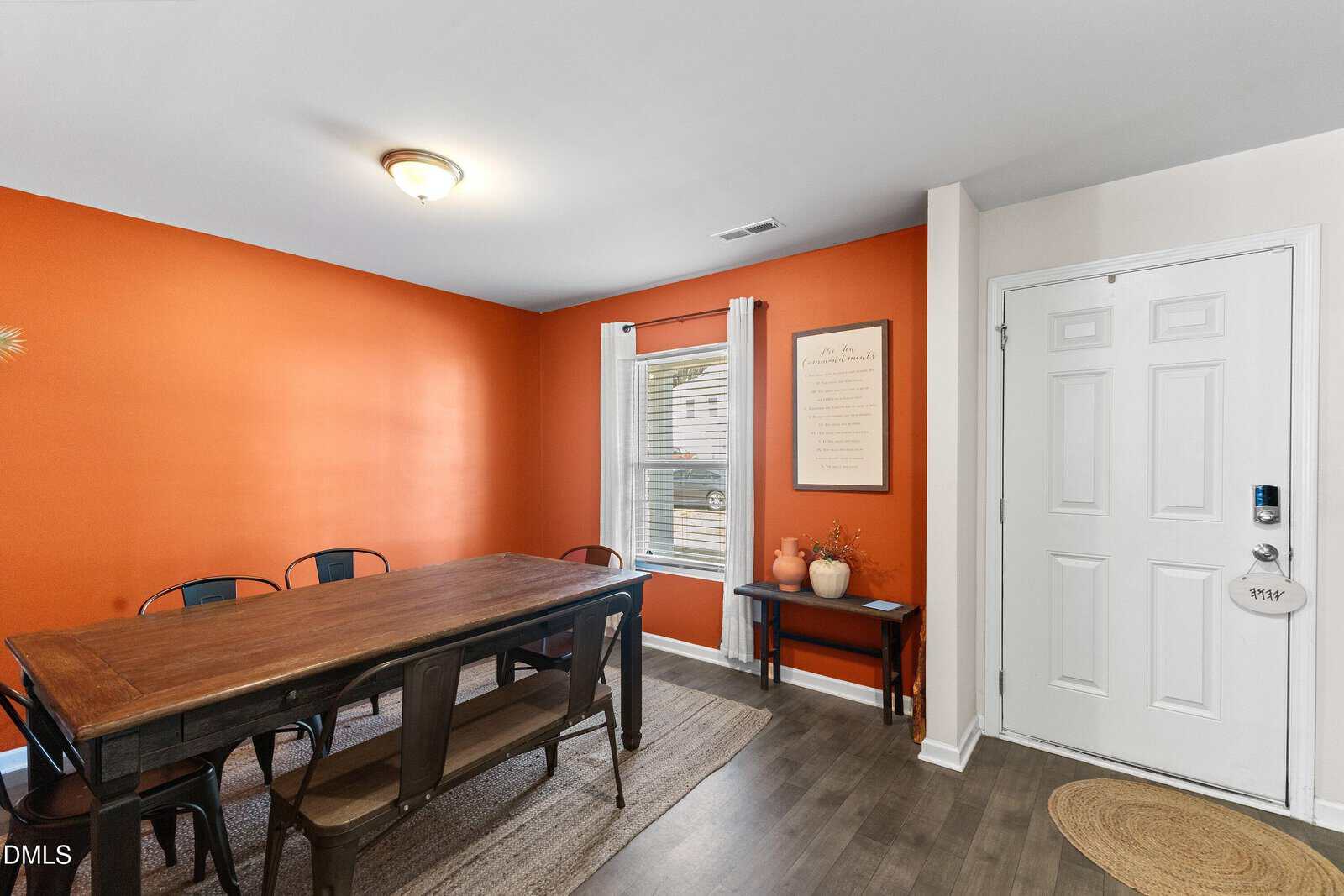 A dining area with an orange accent wall, a wooden table with six metal chairs, a window with white curtains, and a white front door. A small table with decor and a framed sign sits near the entryway.