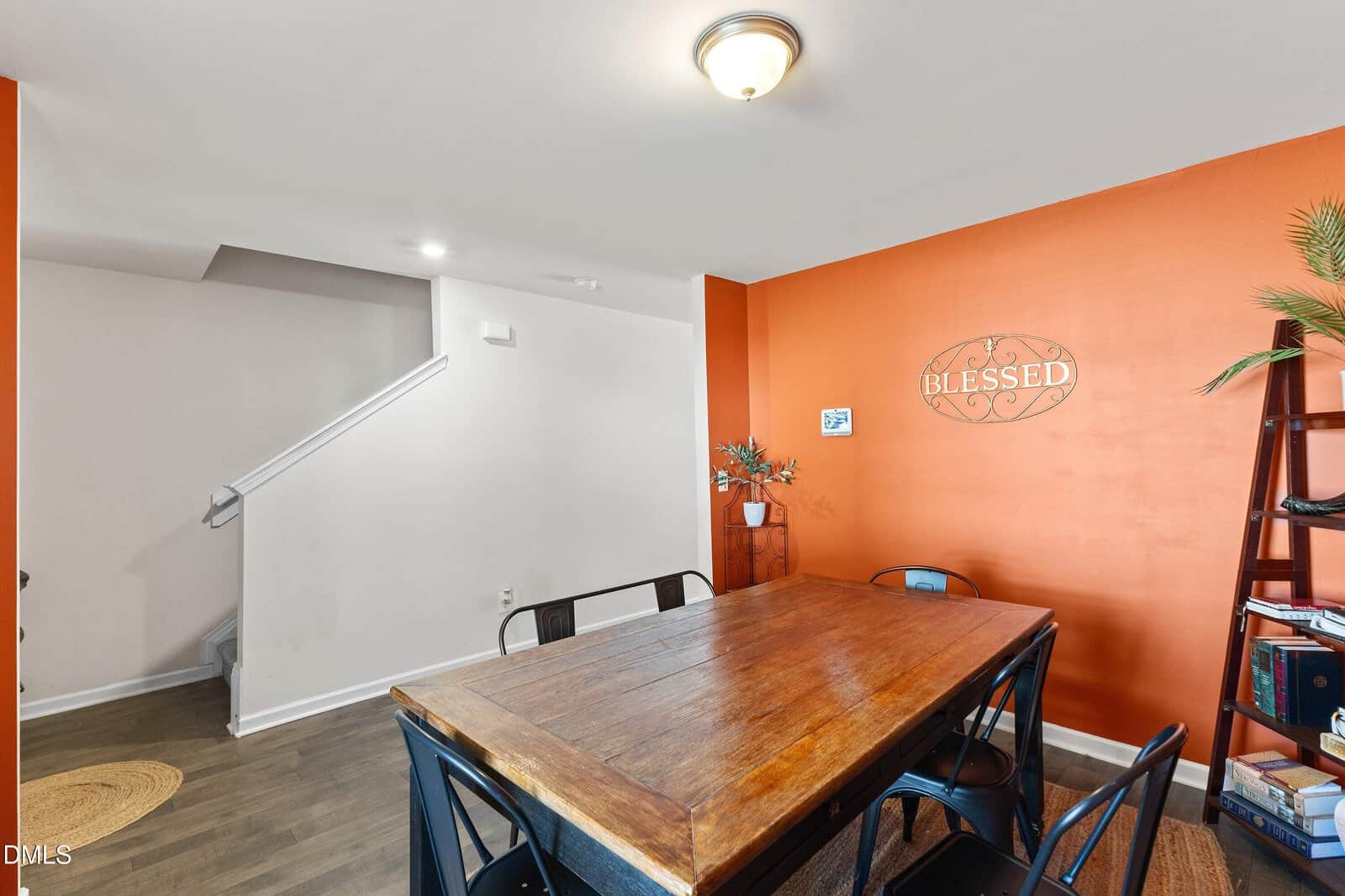 A dining area with a wooden table and six black chairs, an orange accent wall with a "Blessed" sign, a small plant, and a black bookshelf. A staircase with a white railing is visible in the background.