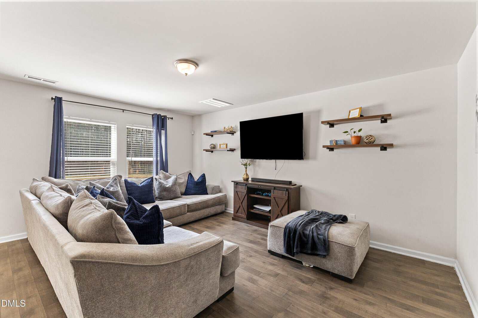 A cozy living room with a large beige sectional sofa, blue and gray pillows, a wall-mounted TV, wooden shelves with decor, an ottoman with a throw blanket, and windows with blue curtains letting in natural light.