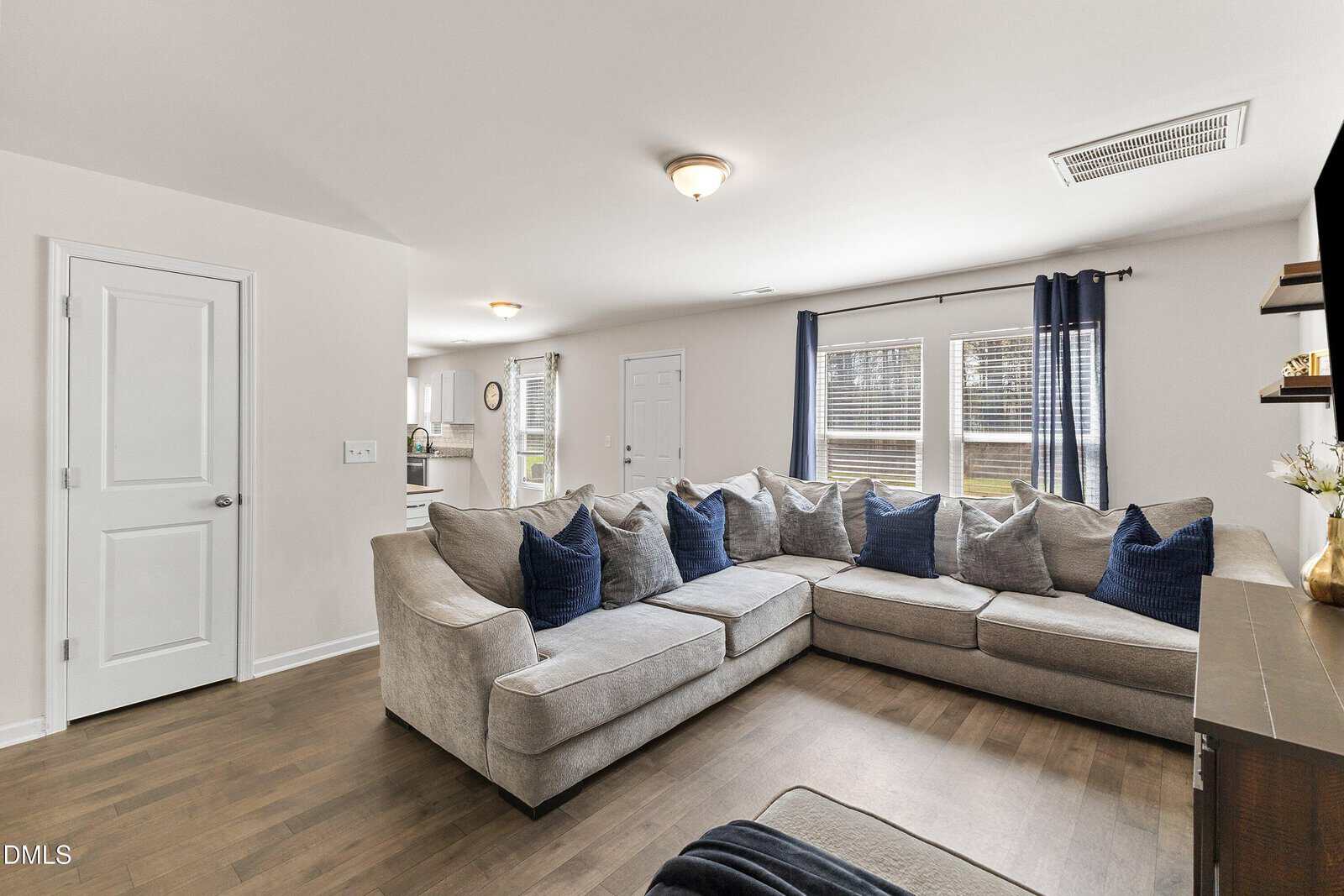 A bright living room with a large L-shaped beige sectional sofa, blue and gray pillows, wooden floors, two windows with blue curtains, and a view into the kitchen area.