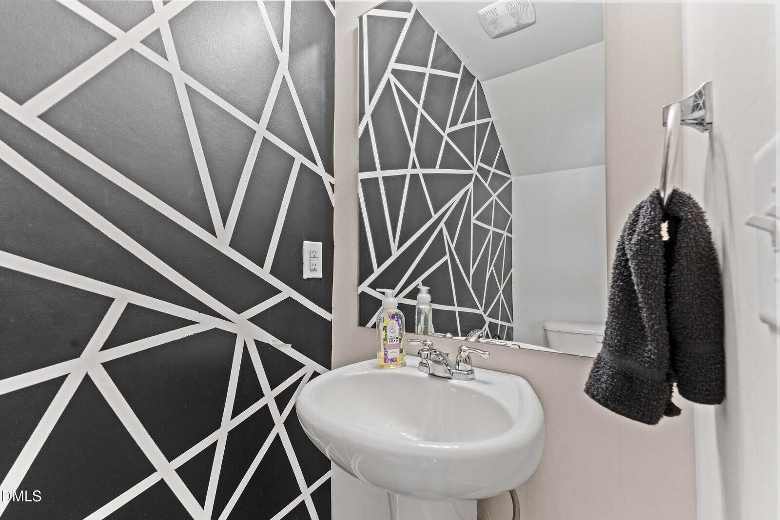 Modern bathroom with a white pedestal sink, silver faucet, soap, towel on a ring, mirror, and a bold black wall with white geometric lines as decor.