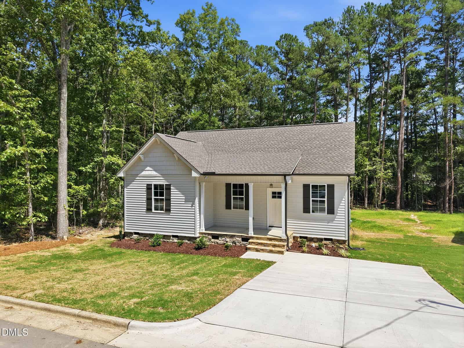 A single-story white house at 137 Seaman Street in Oxford features a front porch, gray roof, and concrete driveway. Nestled next to a wooded area with tall trees under a clear blue sky, the lawn is partially shaded, with some planted shrubs near the foundation.
