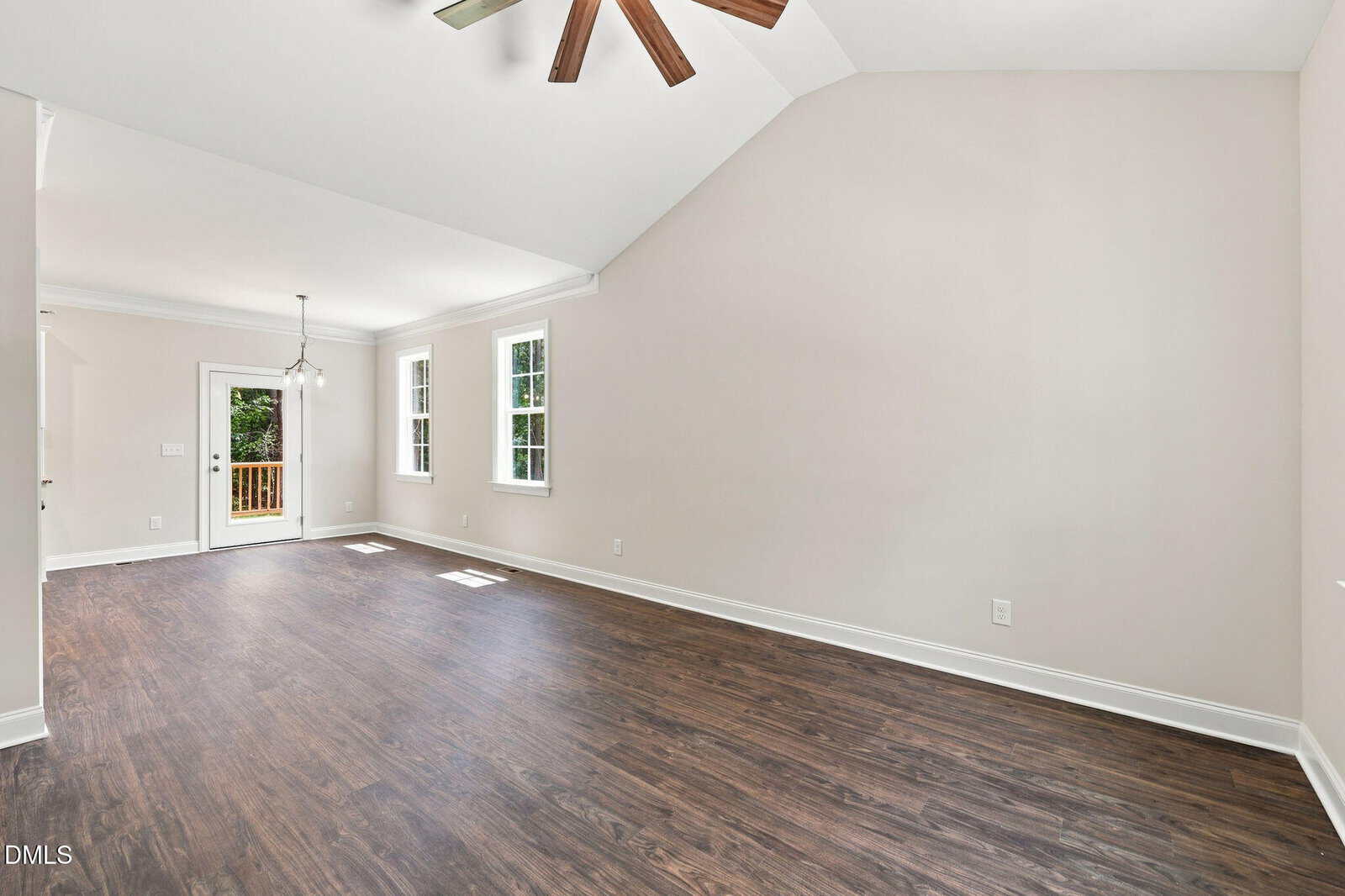 Empty room with white walls, dark wood flooring, vaulted ceiling, ceiling fan, and two large windows. The space extends into a dining area with a light fixture and a glass door leading outside.