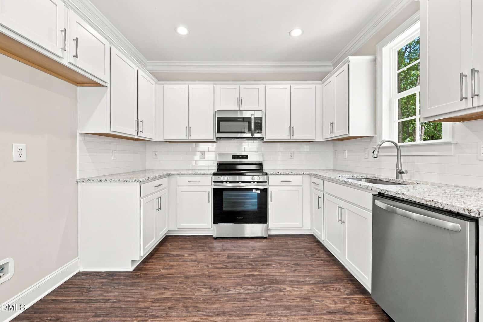 Modern kitchen with white cabinets, granite countertops, stainless steel appliances including oven, microwave, and dishwasher, a single window above the sink, and dark wood flooring. Walls are light beige with white backsplash tiles.