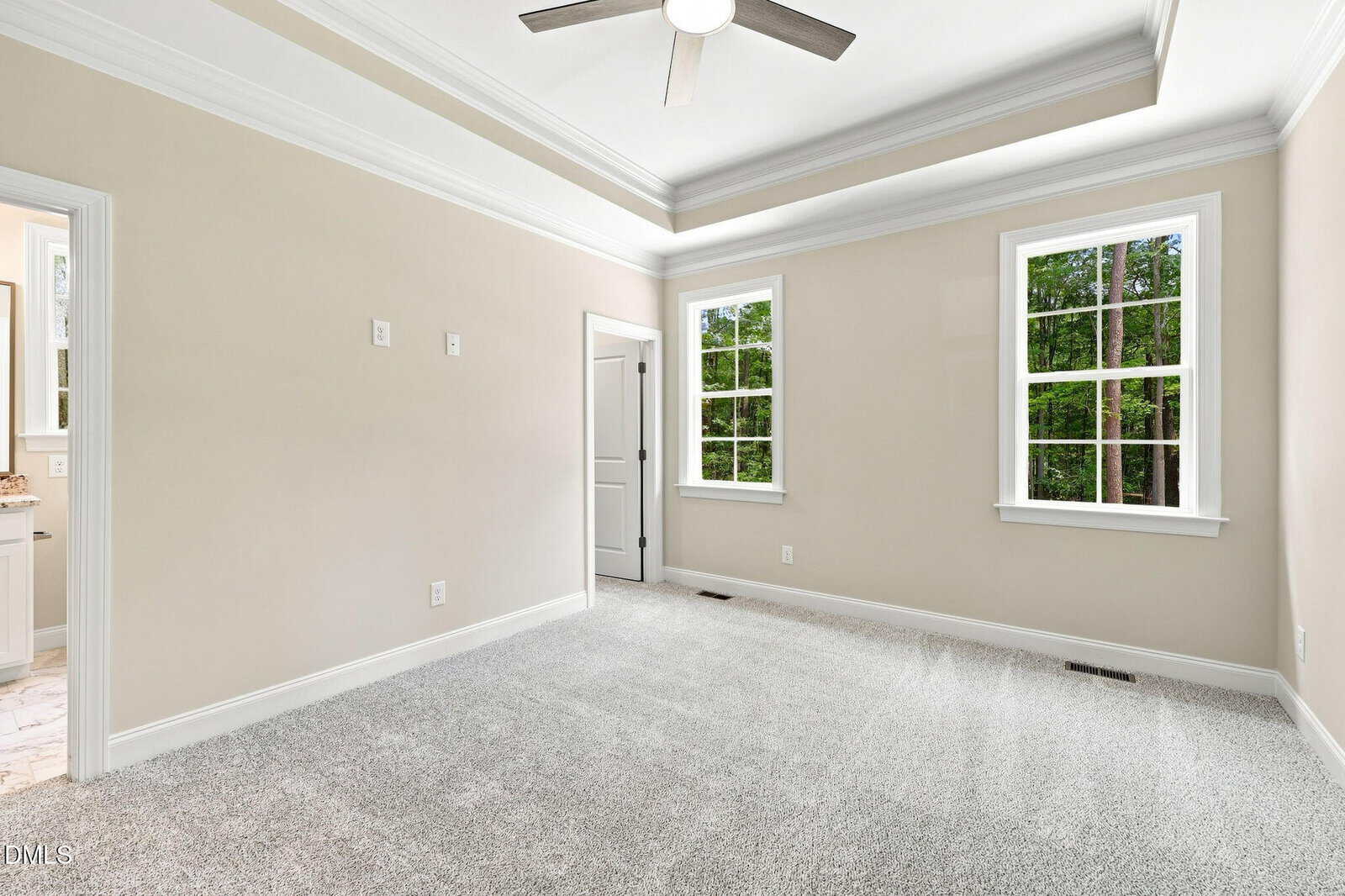 Bright, empty room with beige walls, gray carpet, tray ceiling with fan, two windows showing trees outside, and a white door leading to another space.