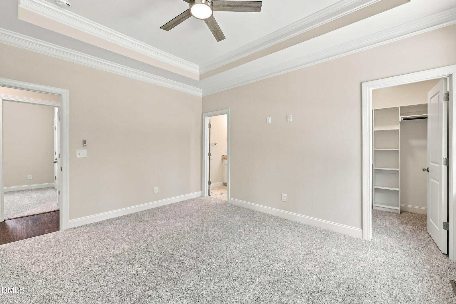 A bright, empty bedroom with beige walls, light carpet, a ceiling fan, two doorways, and a walk-in closet with built-in shelves.