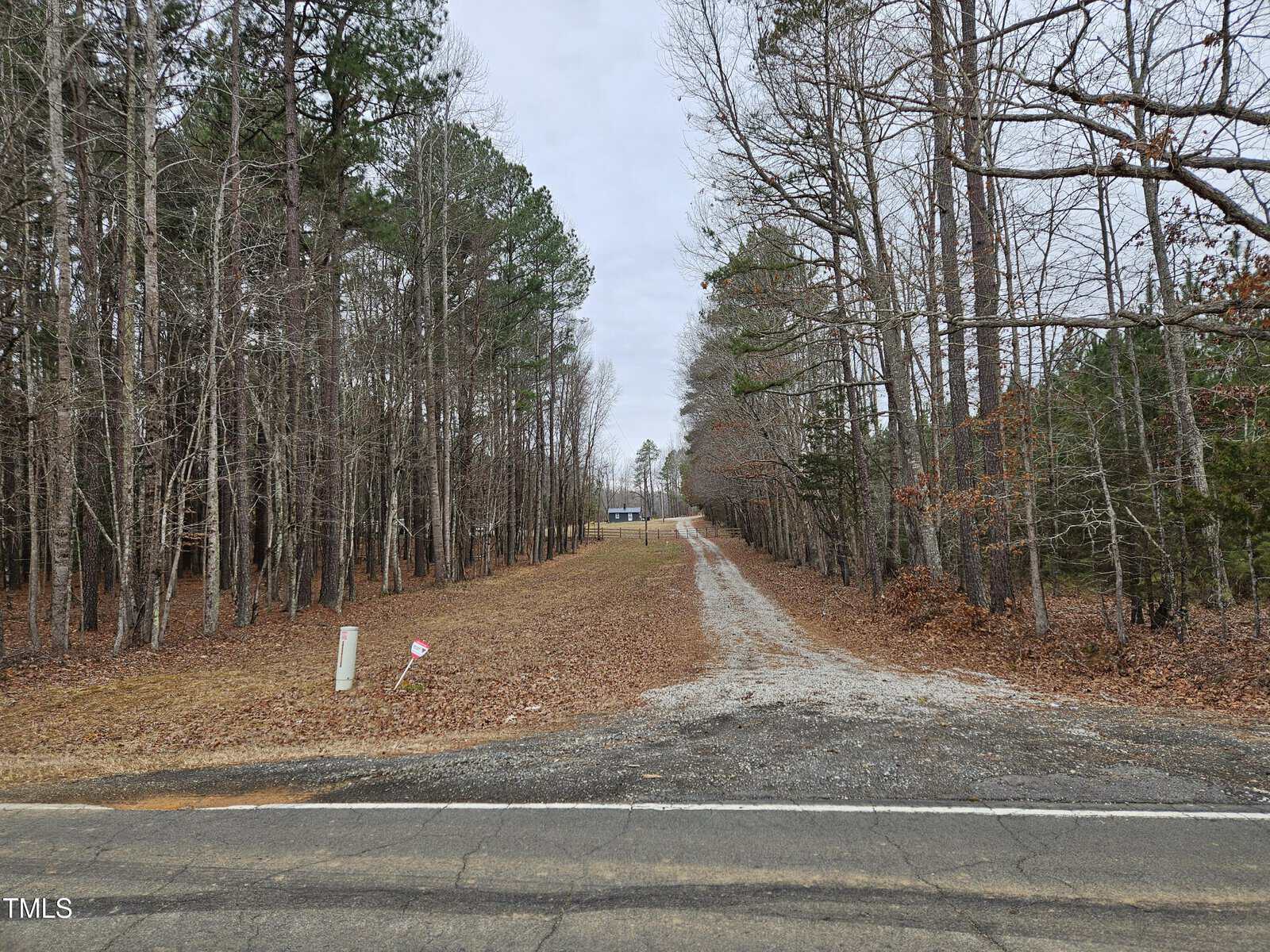 A gravel driveway leads from a paved road through a clearing between tall trees, ending at a distant house. The trees are mostly leafless, and a small utility marker is visible near the road. The sky is overcast.
