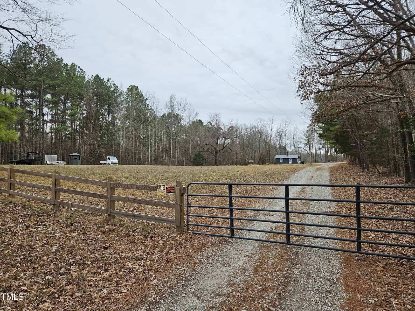 A closed metal gate blocks a gravel driveway leading to an open field with several vehicles and a small building. The area is surrounded by trees under a cloudy sky.