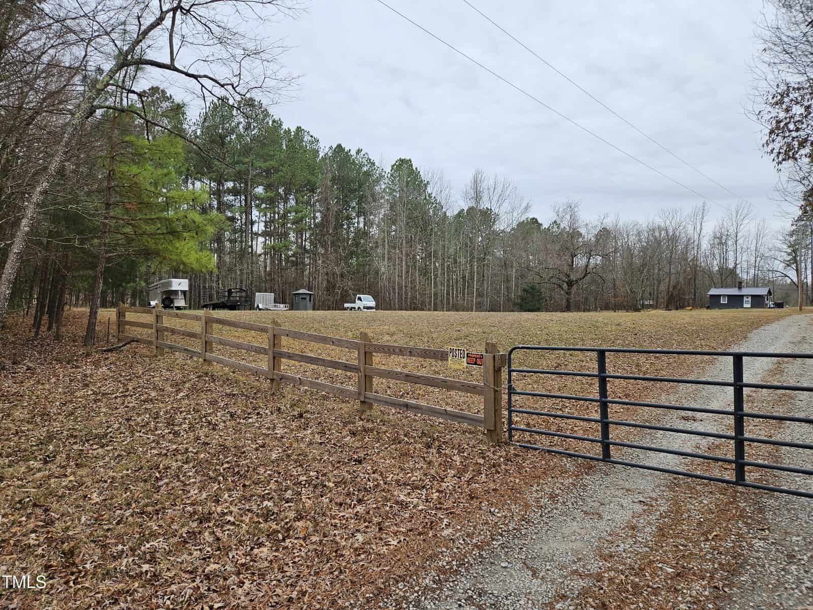 A wooden fence with a black metal gate borders a gravel driveway leading to a grassy field with parked vehicles, trees in the background, and a small building on the right under a cloudy sky.