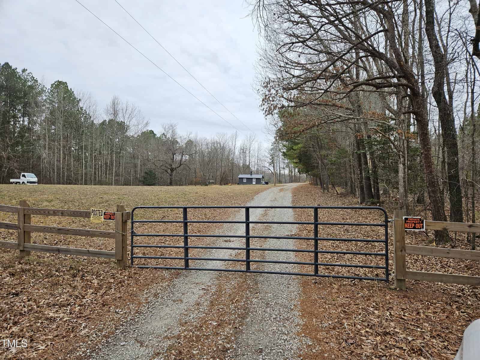 A gravel driveway is gated by a metal fence with "No Trespassing" and "Posted Keep Out" signs; the drive leads through a grassy field to a small house surrounded by trees with a white van parked to the left.