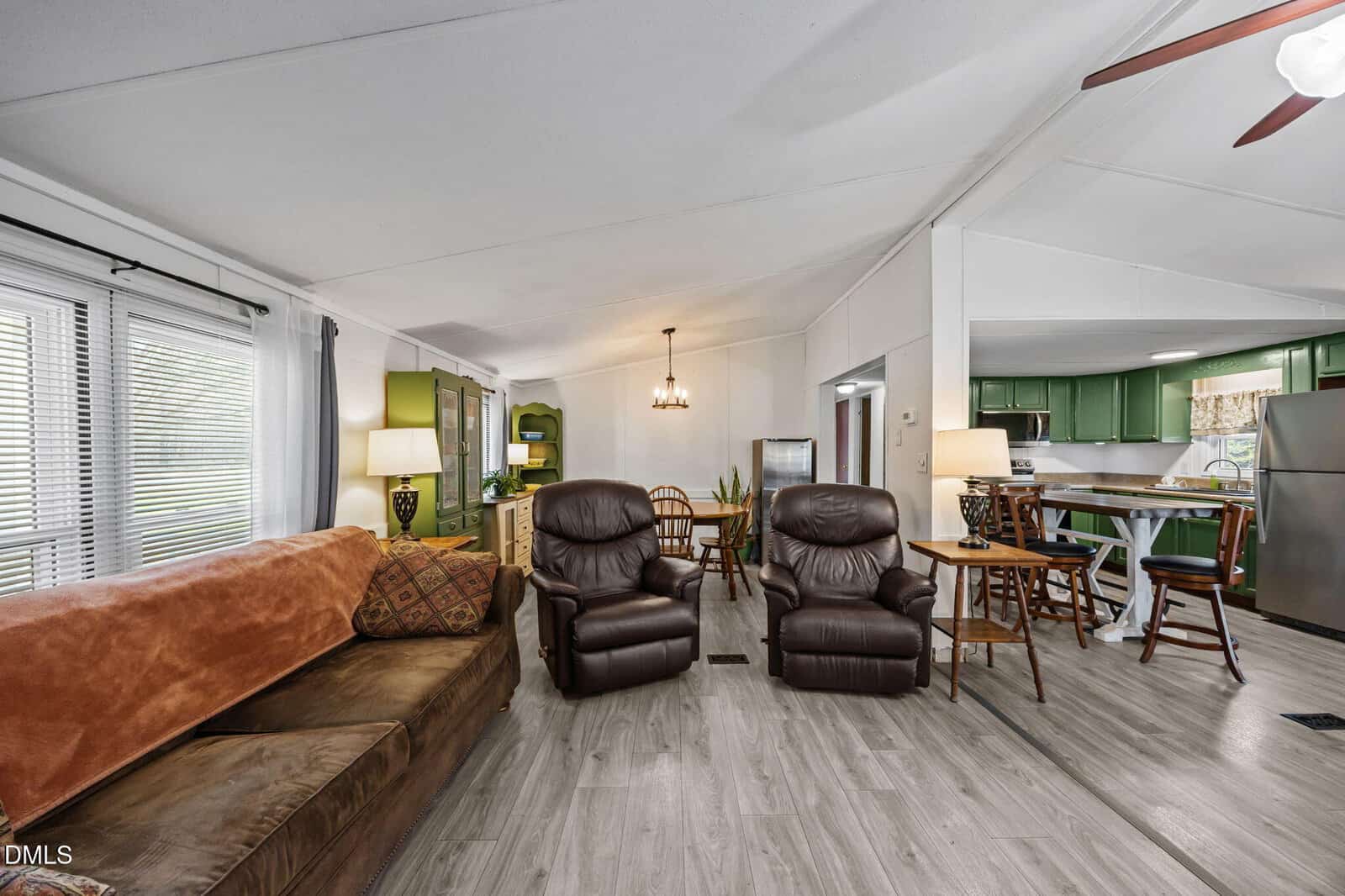 A cozy living room with a brown sofa, two recliners, lamps, dining table, and a kitchen with green cabinets and stainless appliances. Wood flooring and large windows add natural light.