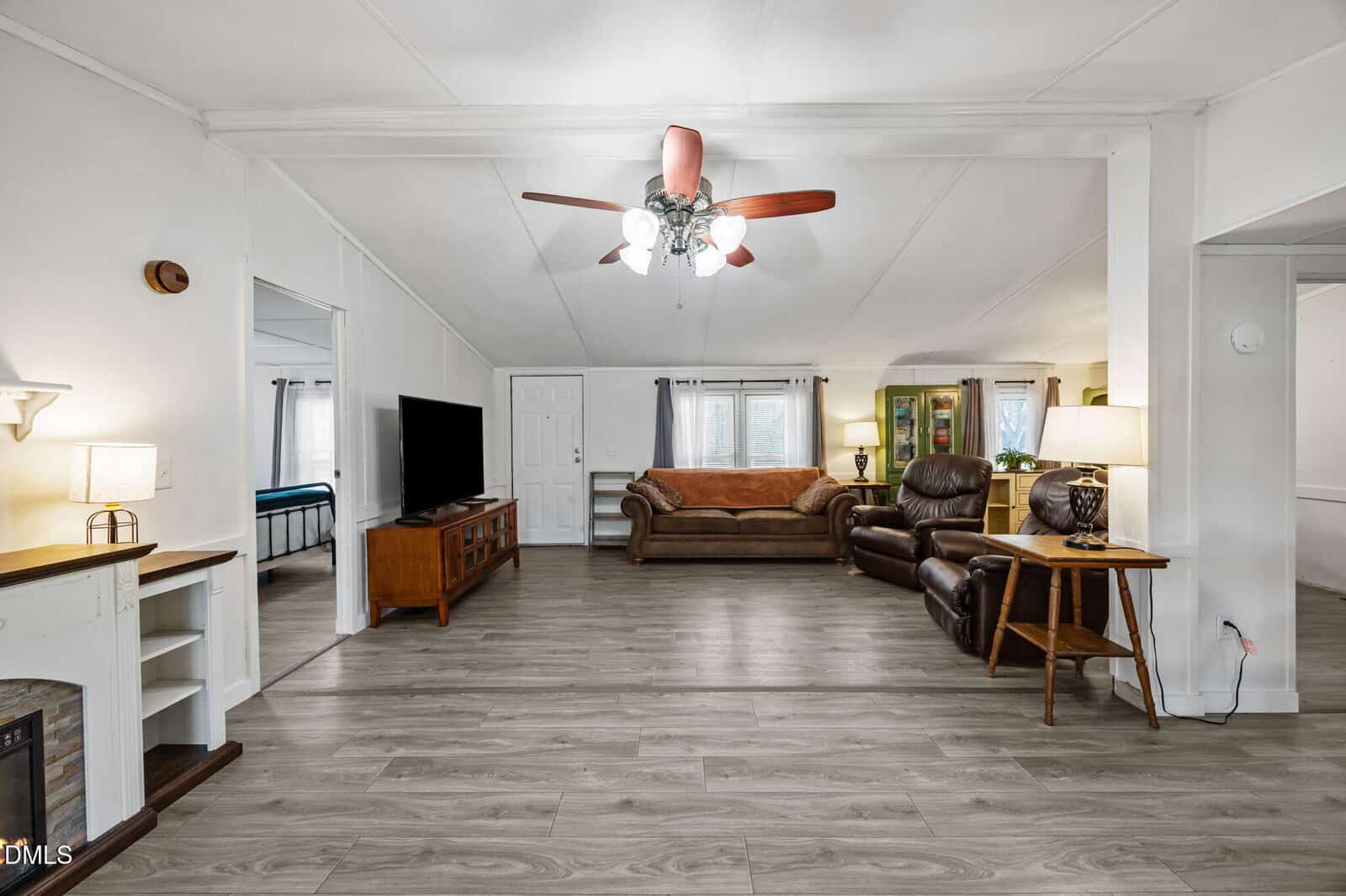 Spacious living room with wood floors, ceiling fan, brown sofa, two recliners, TV on stand, and large window. Open layout with visible adjoining rooms and neutral wall colors.