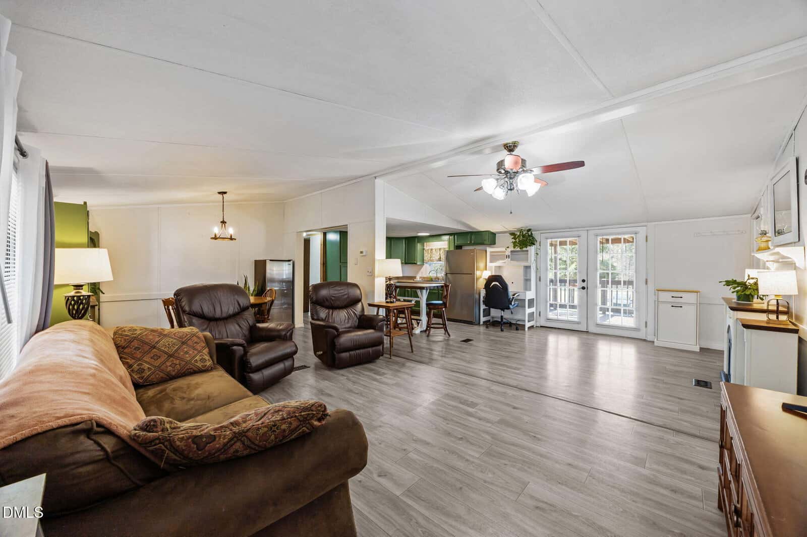 A living room with a brown sofa, two recliners, a ceiling fan, and wood-look flooring. The space opens to a dining area, kitchen with green accents, and glass doors leading outside. A desk is situated near the kitchen.