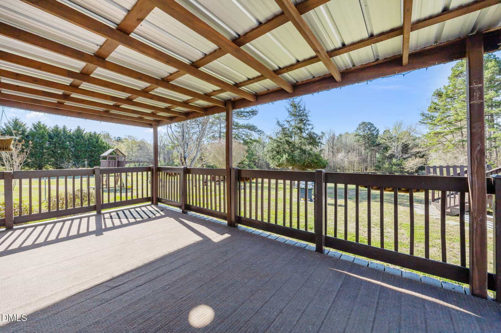 A covered wooden deck with a slatted roof and railing overlooks a grassy yard with trees in the background. The area is well-lit with natural sunlight.