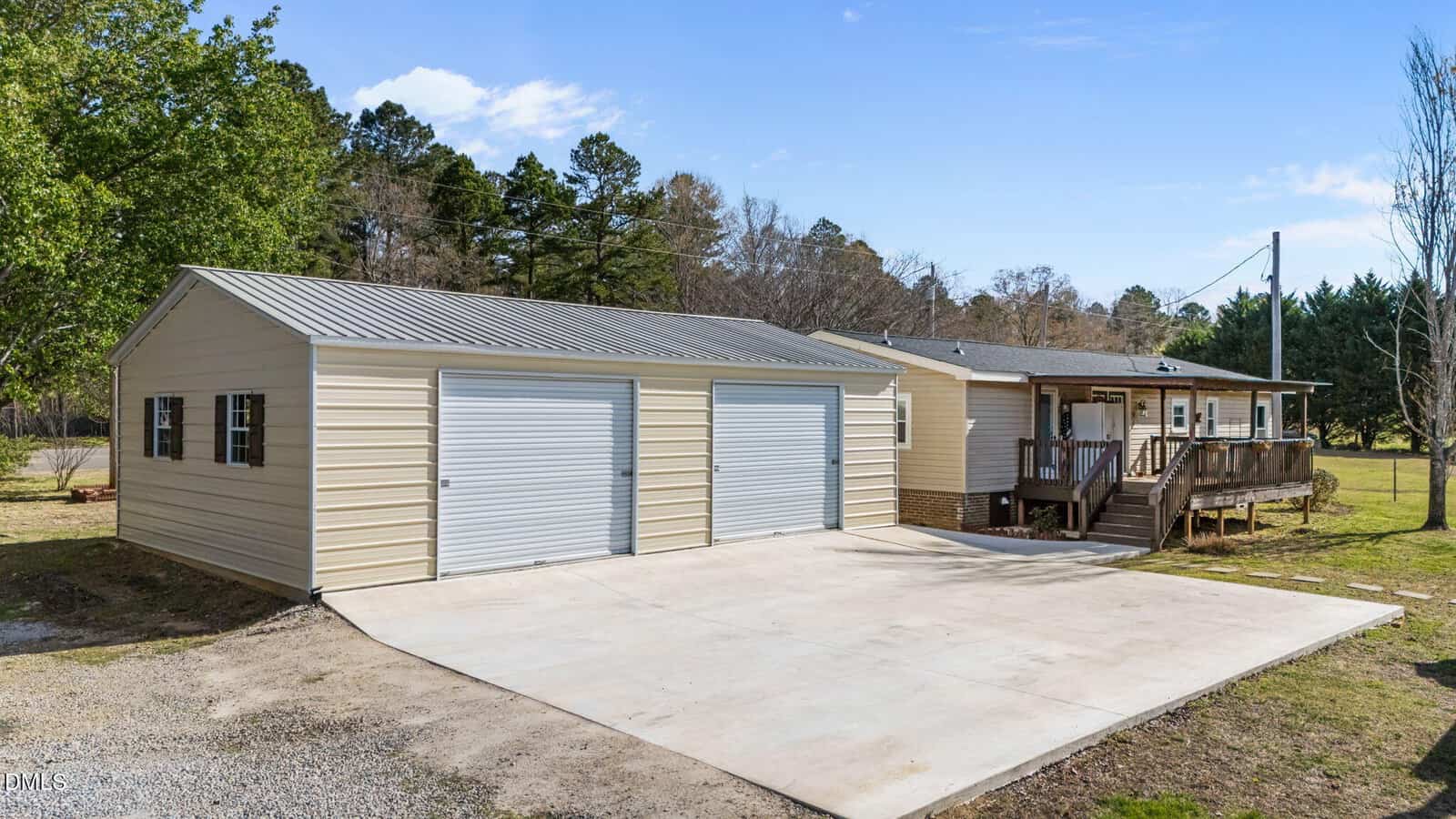 A beige single-story house with a metal roof, attached wooden porch, and a three-car garage with white roll-up doors, set on a concrete driveway amid green trees and grass under a blue sky.