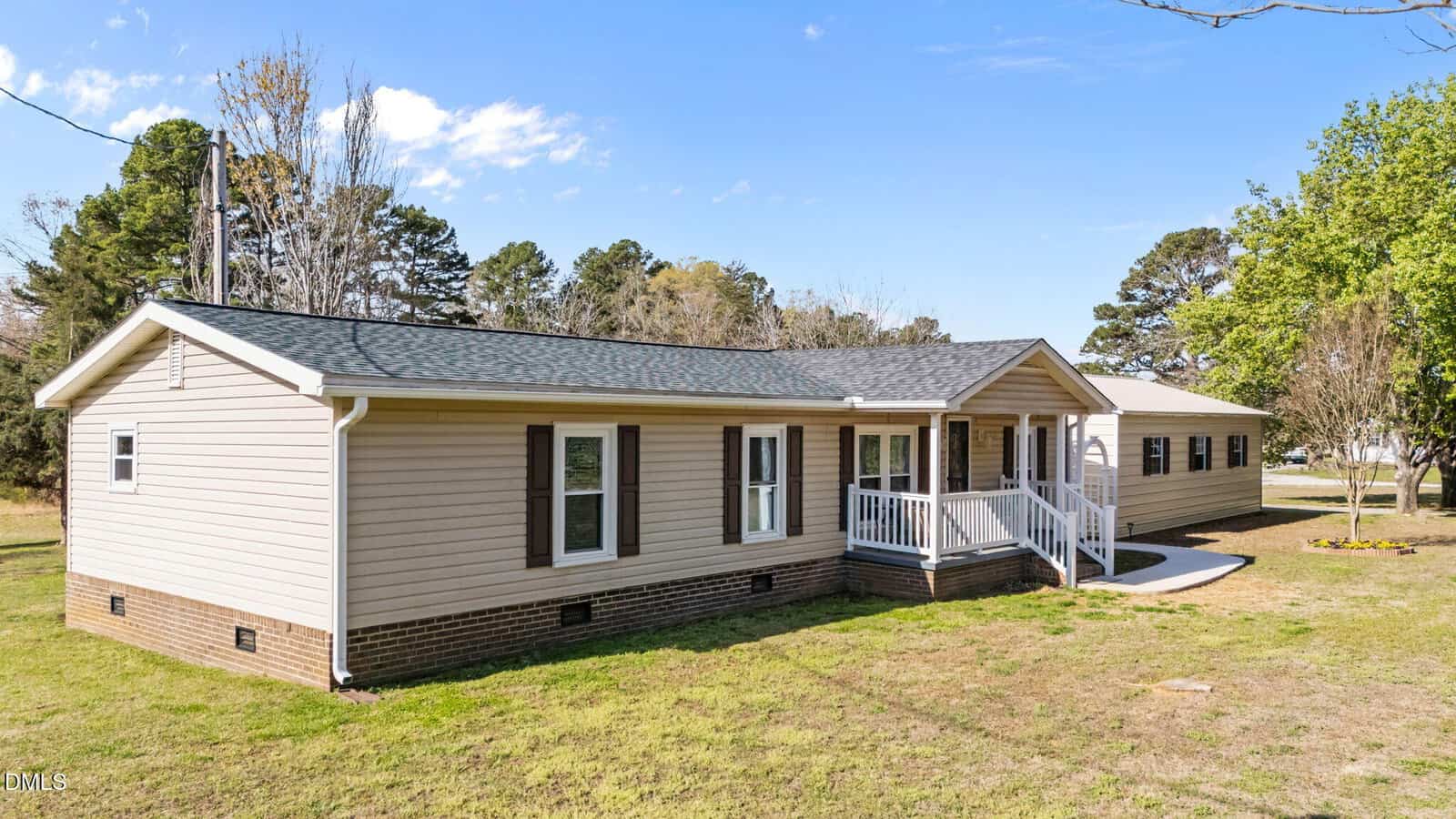A single-story beige manufactured home with white trim, a covered front porch with white railings, black shutters, and a brick foundation sits on a grassy lawn with trees in the background.