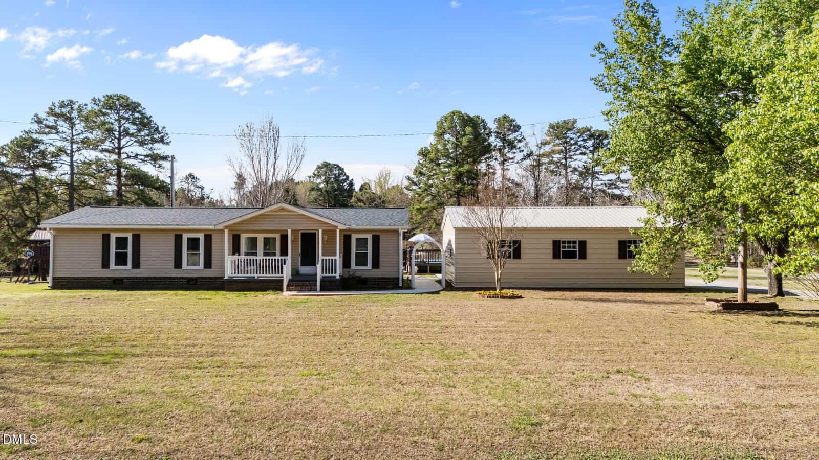 A single-story beige house with a covered front porch sits beside a large detached garage or outbuilding on a spacious, grassy lot with several trees and a clear blue sky.