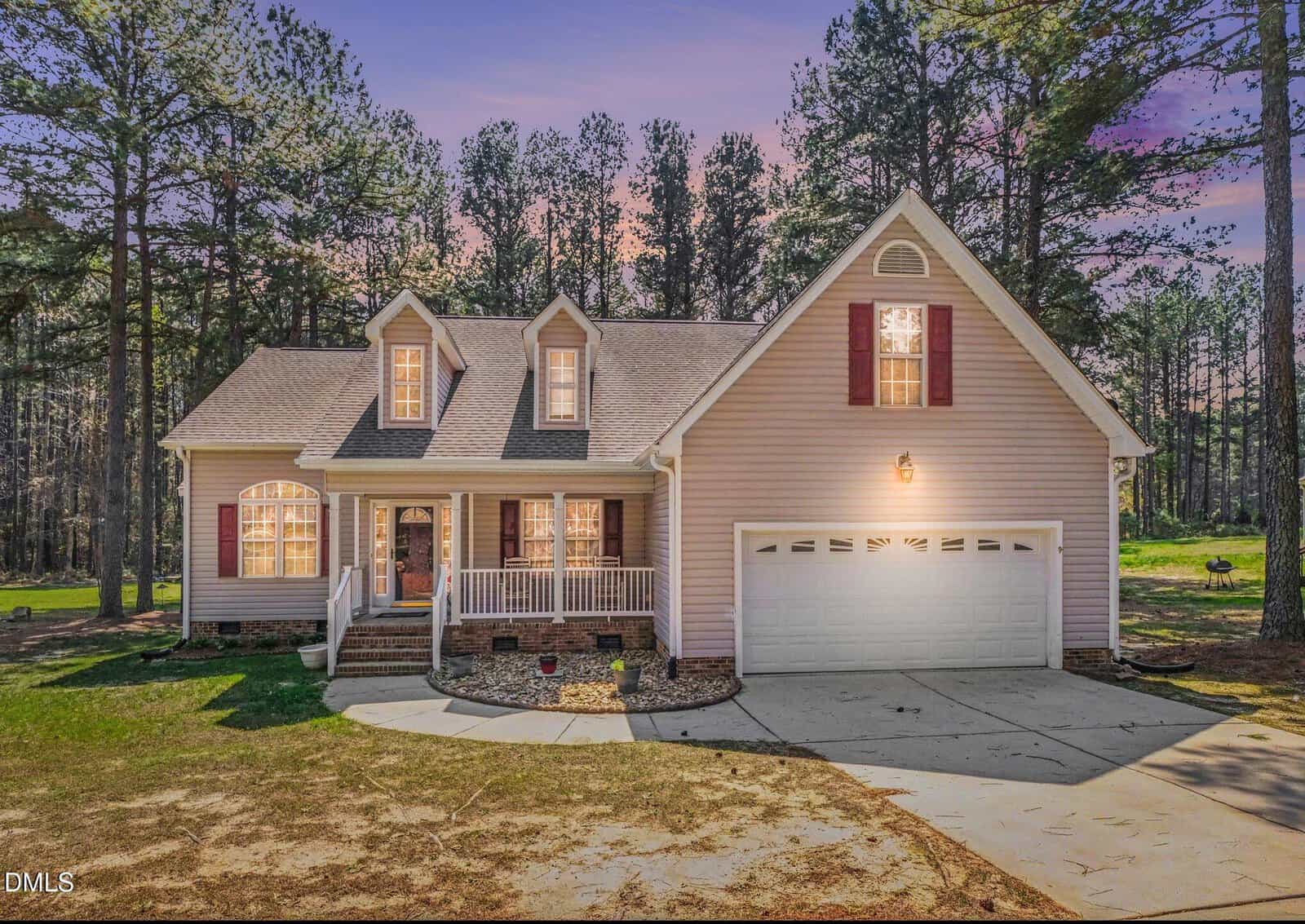 A two-story beige house with a front porch, white railings, red shutters, and a double garage is surrounded by tall trees at sunset. The driveway and front lawn are visible in the foreground.