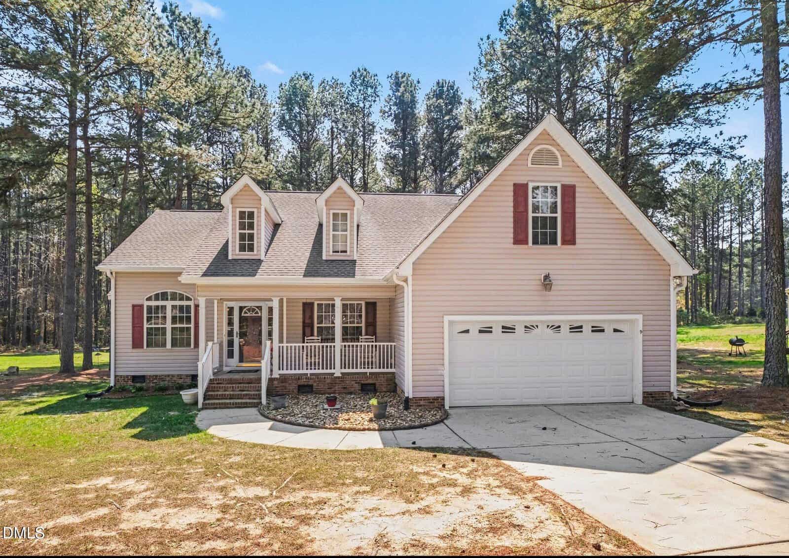A two-story beige house with red shutters, a covered front porch, and a double garage sits on a spacious lot surrounded by tall pine trees under a clear blue sky.