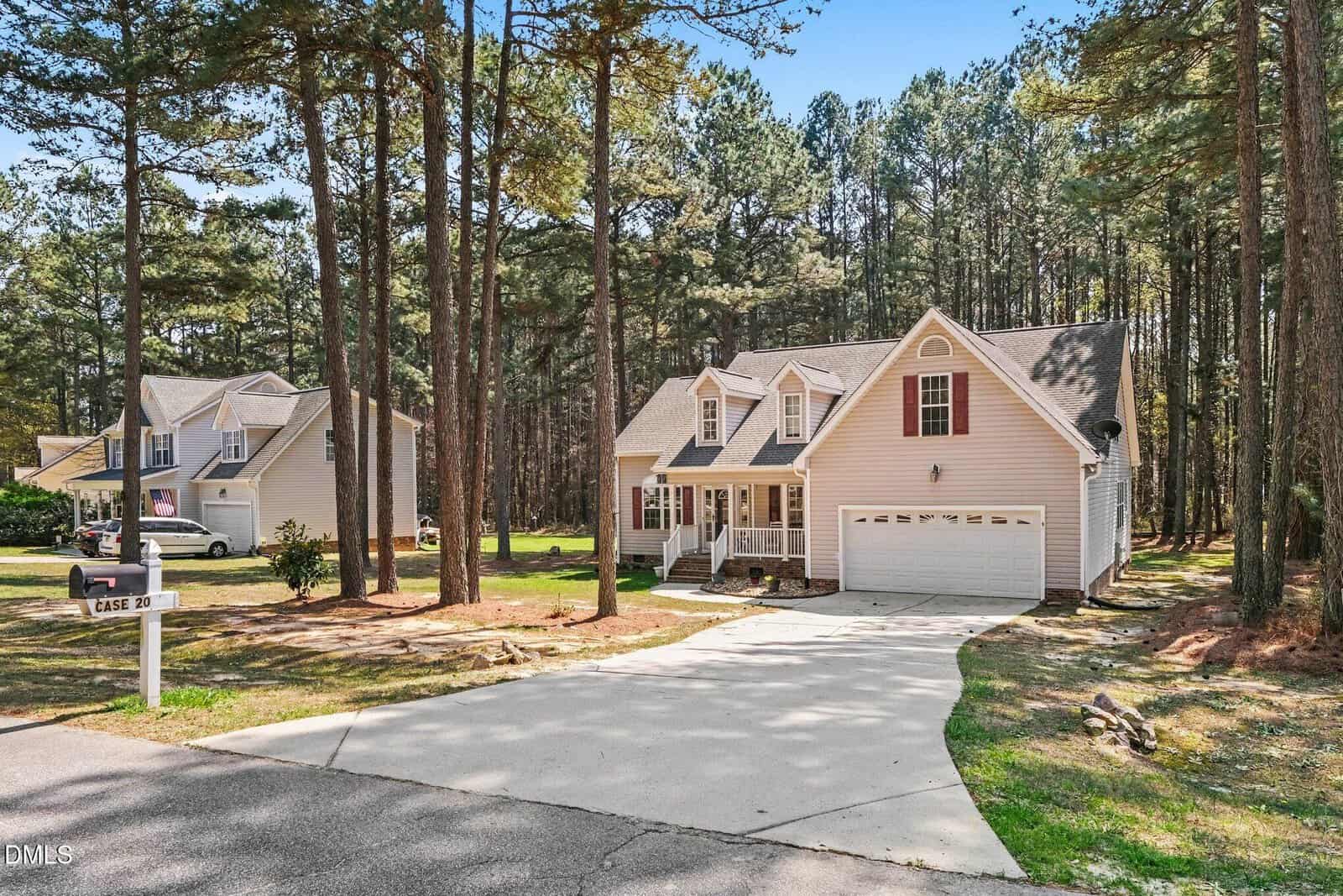 A two-story beige house with white trim and a two-car garage sits among tall pine trees. A white car is parked at the neighboring house, and the driveway curves from the road to the garage. The area is wooded and well-maintained.