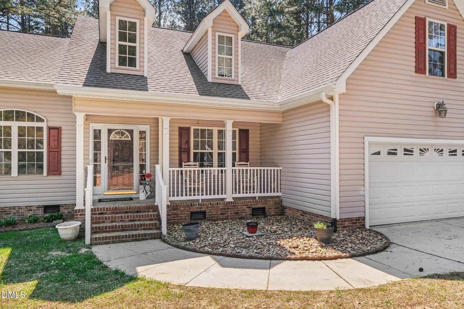A beige house with a small front porch, white railing, brick steps, red shutters, and a white garage door. The front yard has a curved walkway, decorative rocks, and potted plants near the entrance.