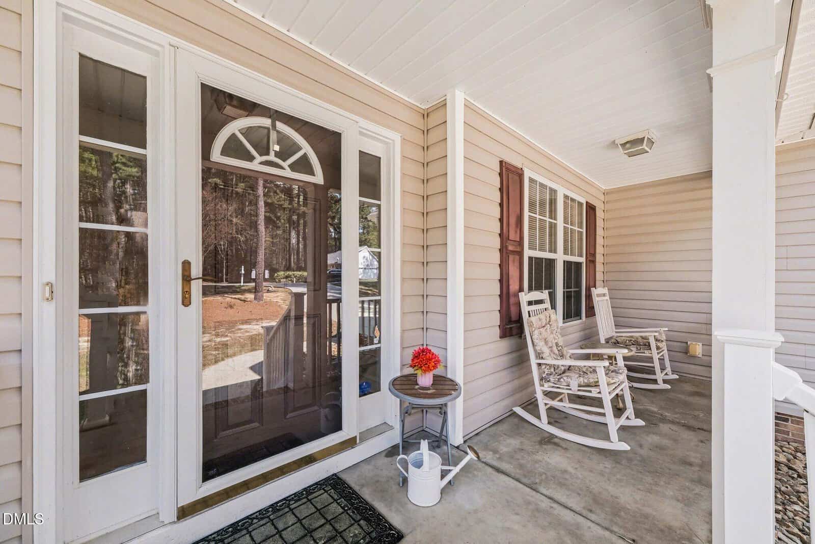 A covered porch with two white rocking chairs, a small round table holding a red flowerpot, a watering can on the ground, beige siding, large windows, and a glass storm door leading inside.