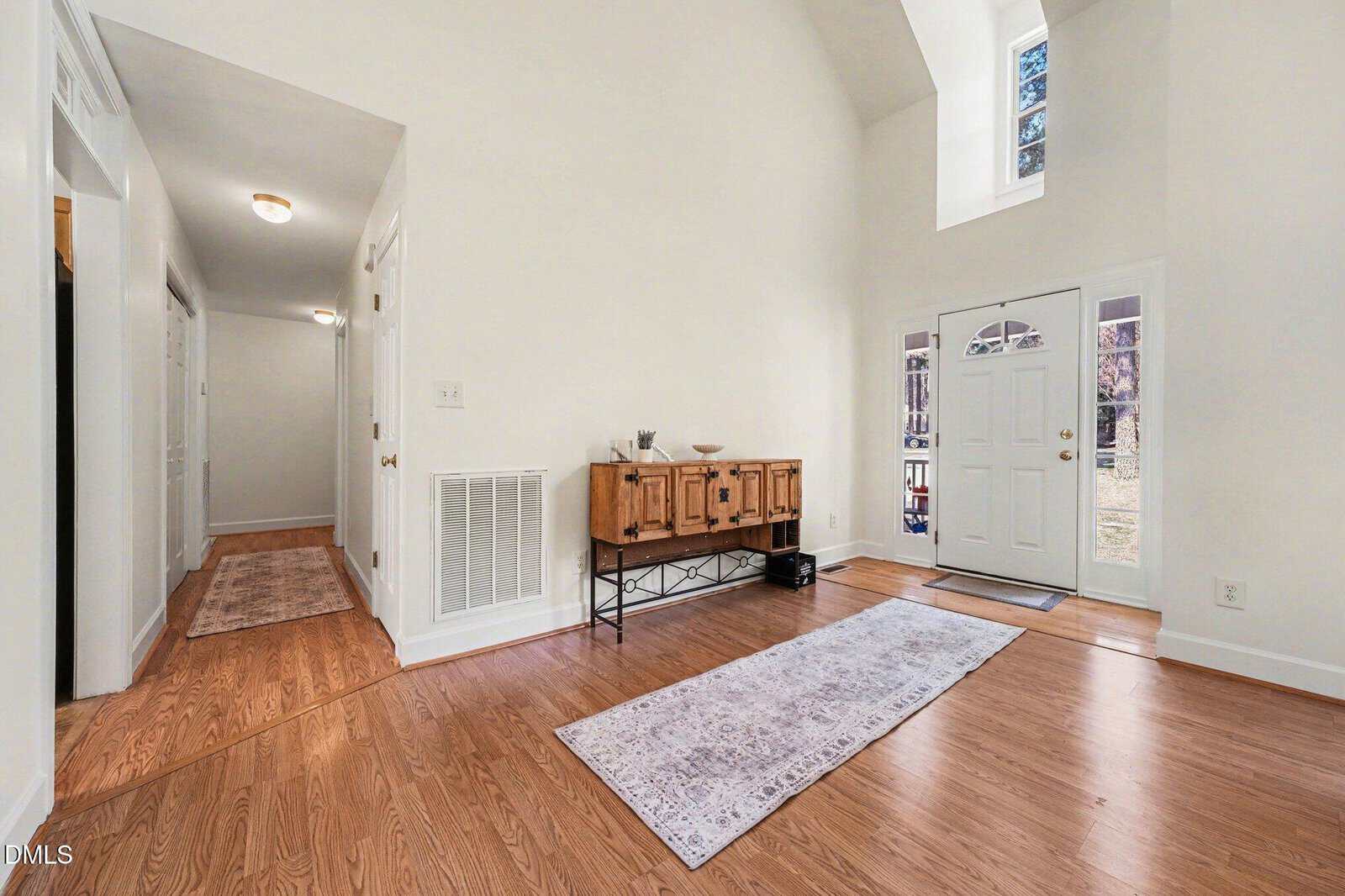 Bright entryway with wood floors, white walls, a tall ceiling, and large windows. A console table and two rugs decorate the space, with a hallway leading to additional rooms on the left.
