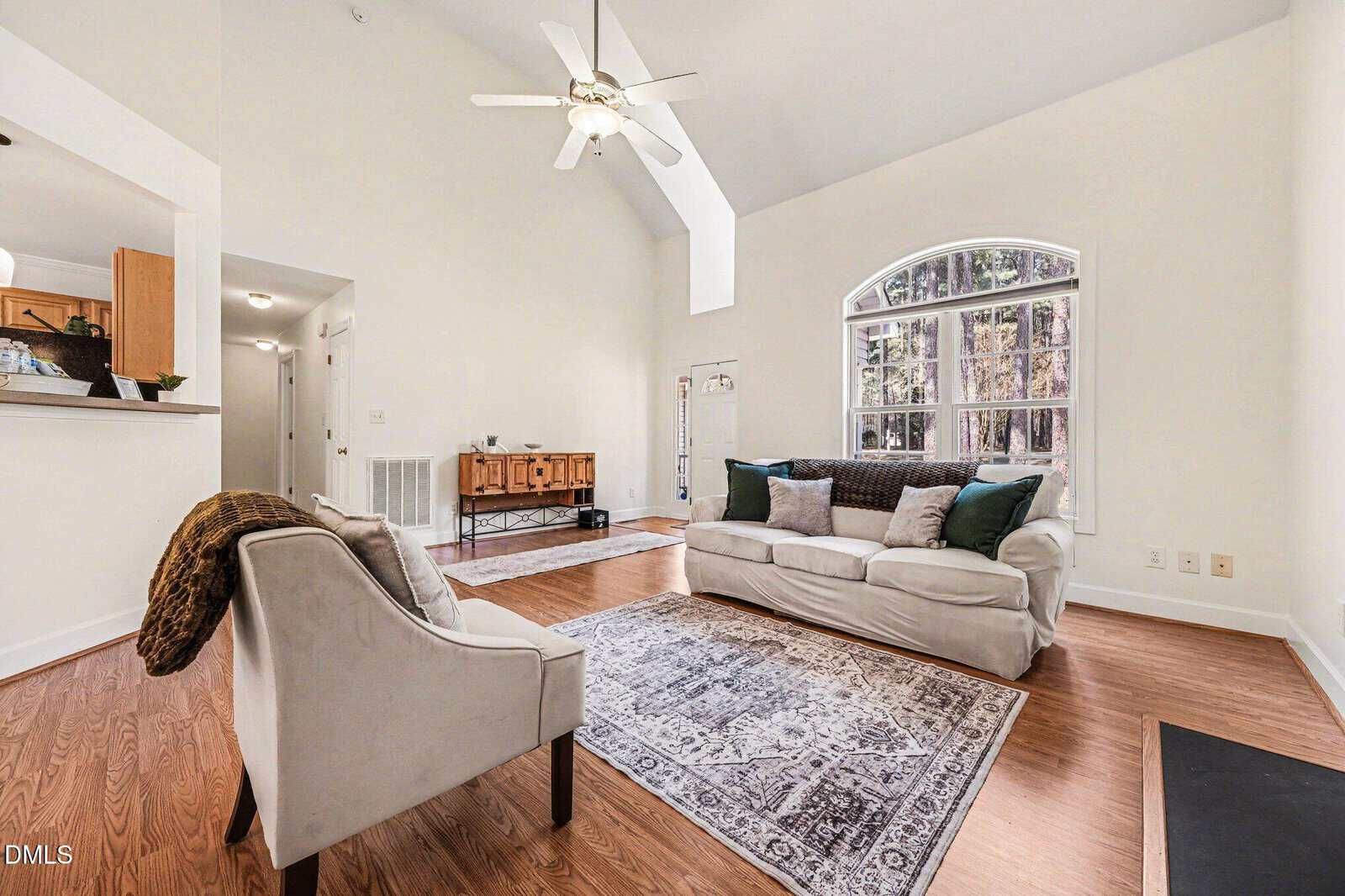 Bright living room with a large arched window, white walls, hardwood floor, beige armchair, white sofa with pillows, patterned rug, ceiling fan, and a wooden sideboard against the back wall.