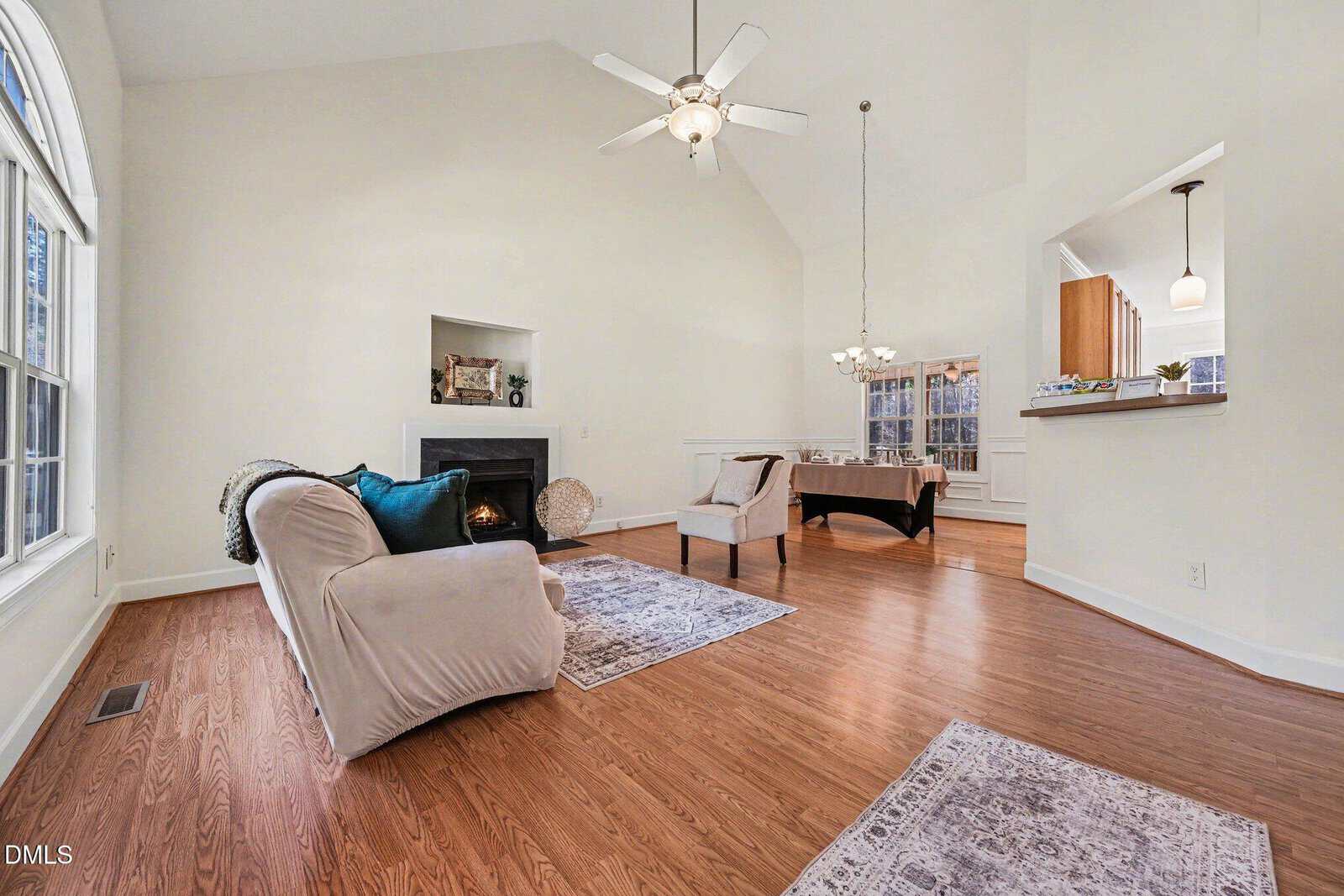 Bright living room with vaulted ceiling, ceiling fan, hardwood floor, fireplace, and neutral-colored furniture. Large window to the left, open dining area and kitchen in the background, modern and airy feel throughout.