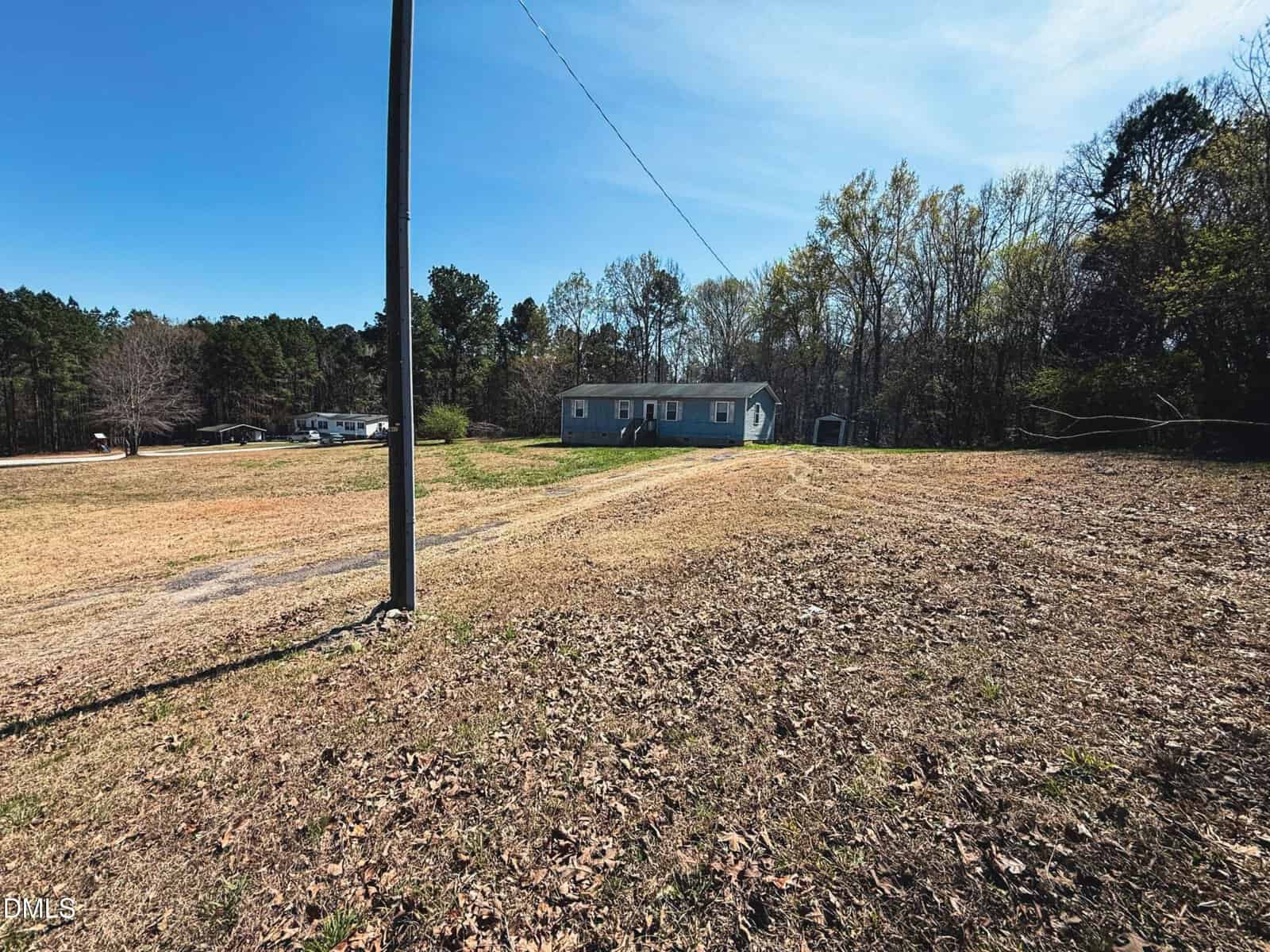 A large, mostly bare grassy lot with a utility pole in the foreground. In the background, there is a light blue one-story house surrounded by trees under a clear blue sky.