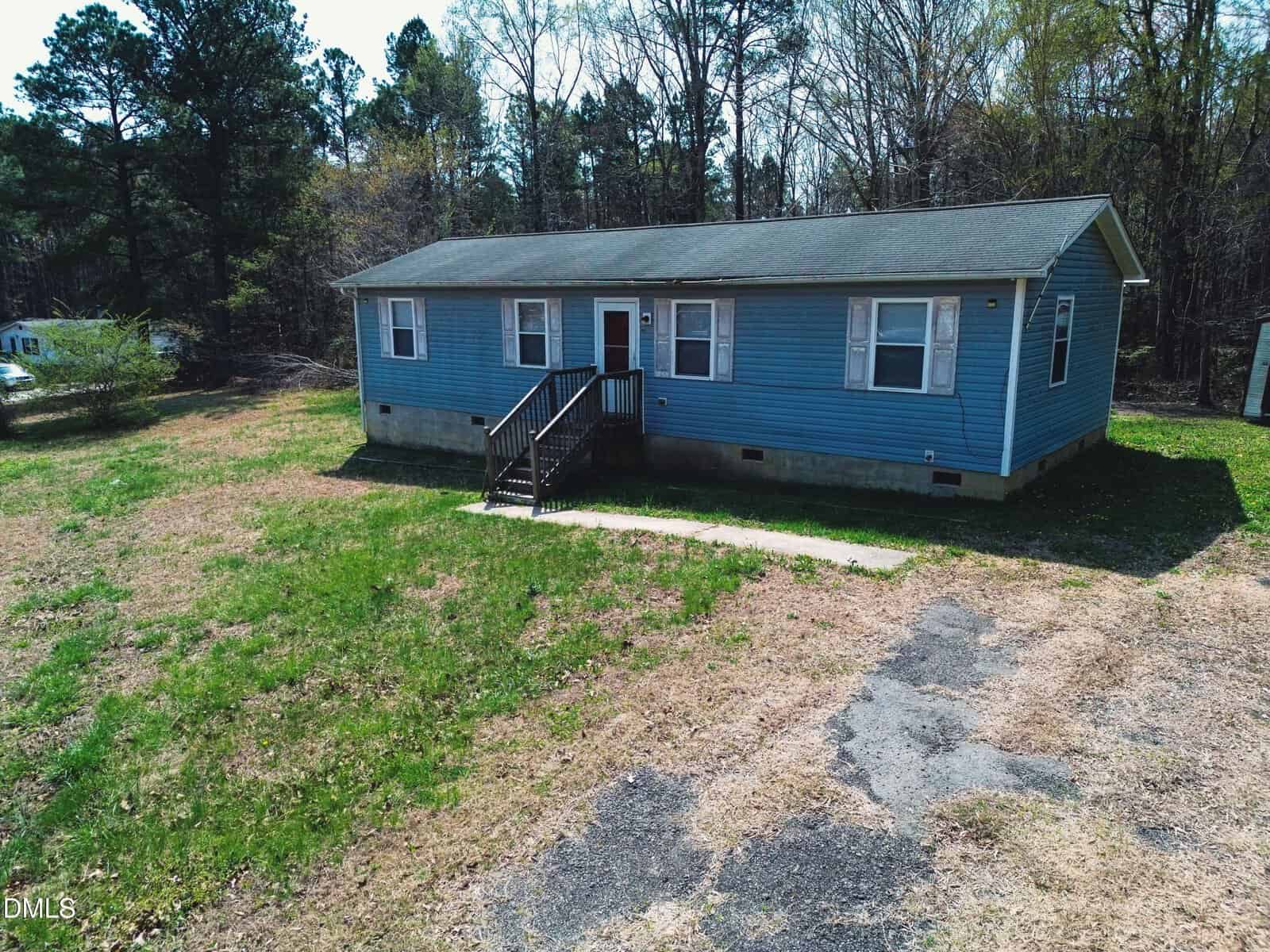 A single-story blue house with white trim, a front porch with steps, and a patchy lawn. The house is surrounded by trees and sits on a slightly raised foundation. There is a partially paved driveway in front.