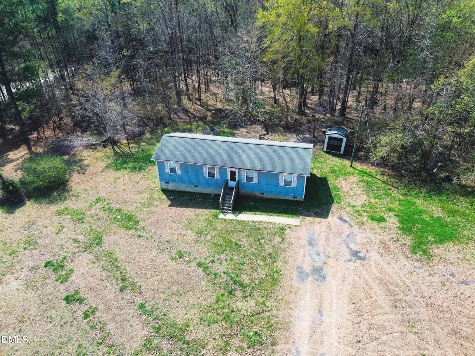 A small blue single-story house with a front porch sits on a patchy lawn near a wooded area. A small shed is visible behind the house. The yard is mostly bare with some grass and trees around the property.