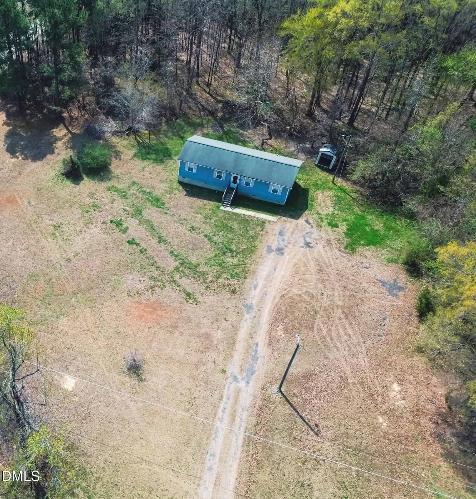 Aerial view of a small blue house with a front porch, surrounded by trees and a dirt driveway, set in a spacious, grassy, rural area.