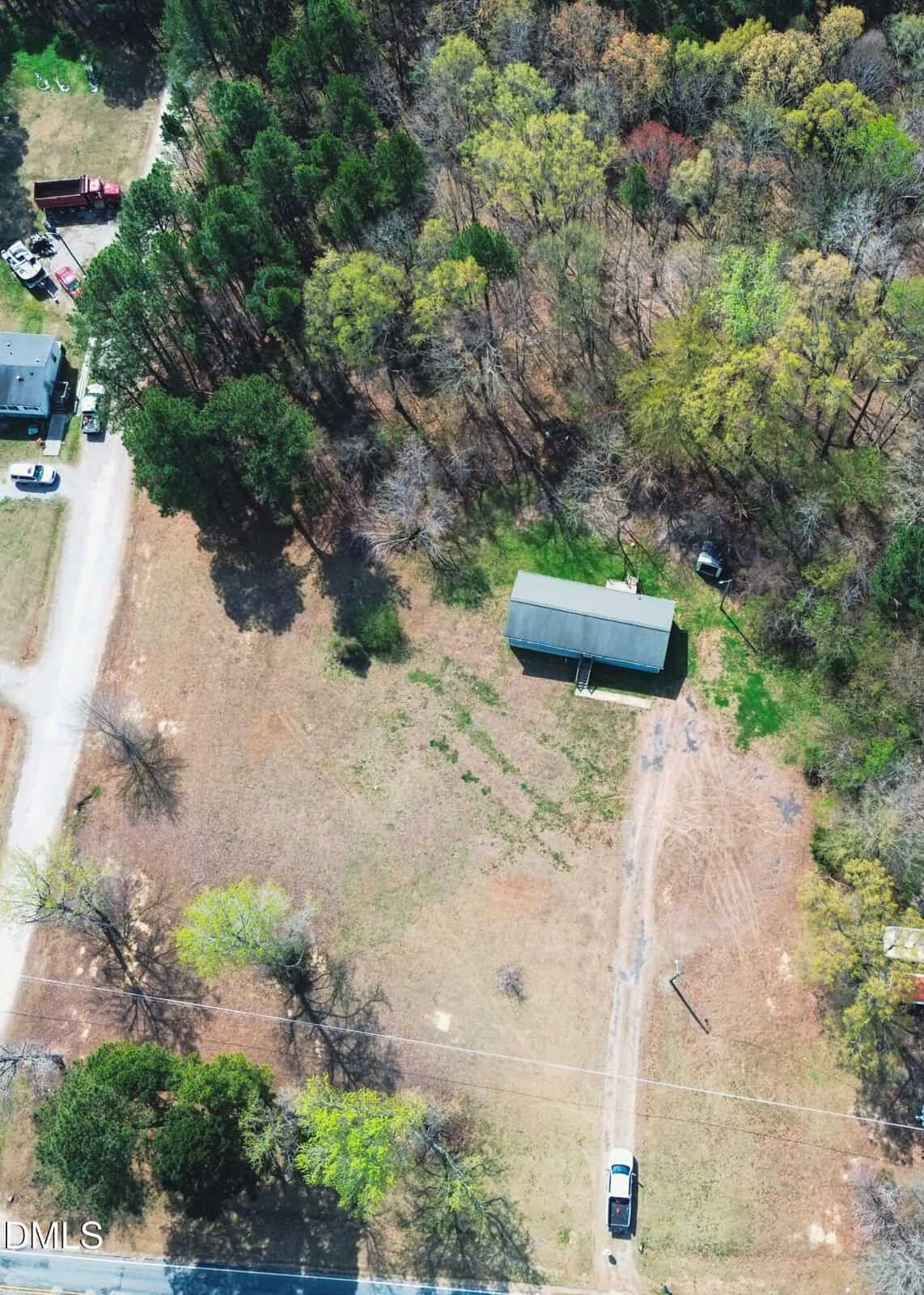 Aerial view of a rural property with a single house, trees around the edges, a dirt driveway, and two vehicles—one near the house, another by the road. Sparse grass and mixed tree coverage are visible.