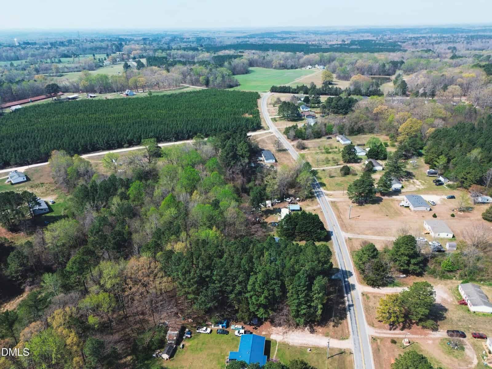 Aerial view of a rural area with scattered houses, winding roads, green fields, and dense forests under a clear sky.