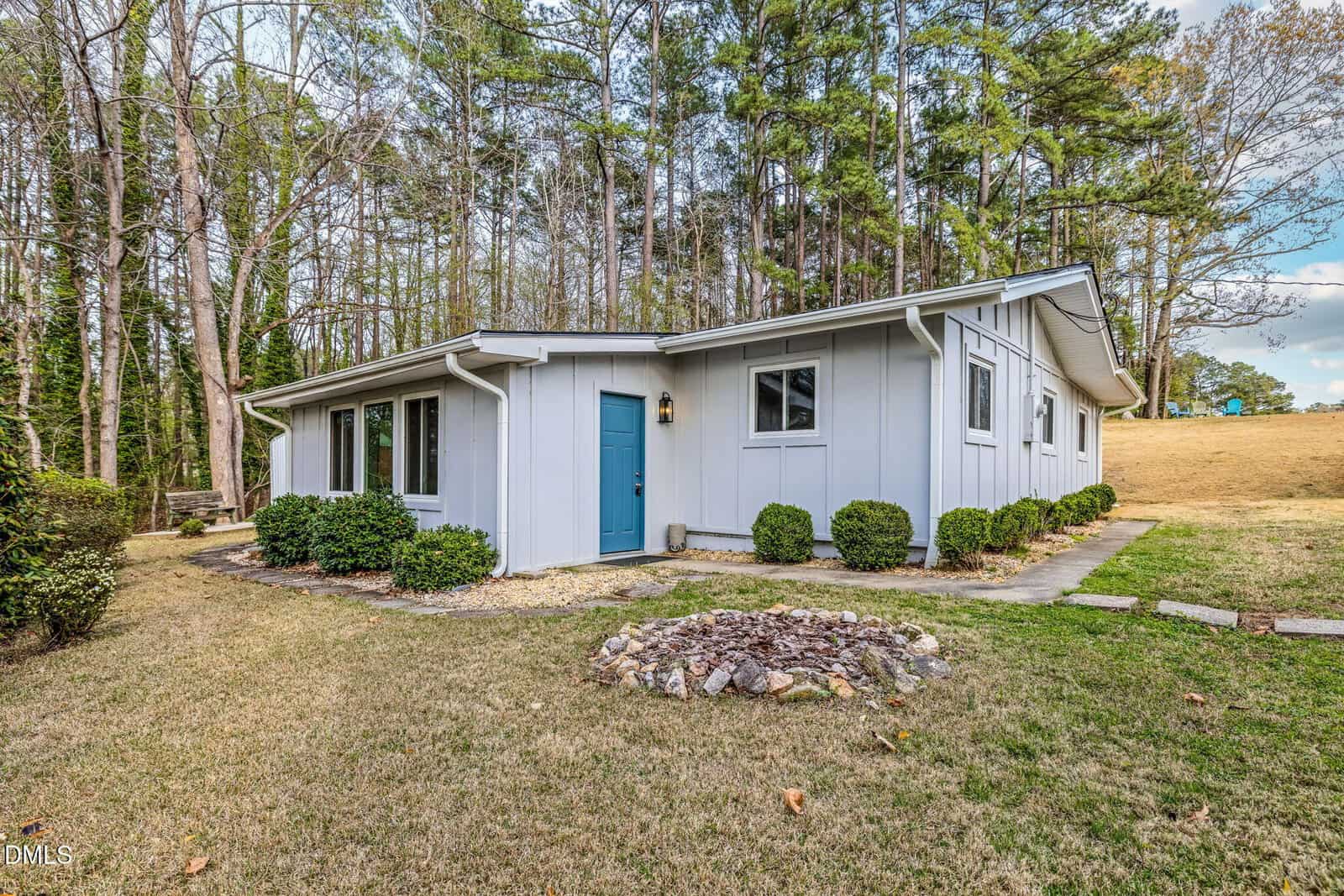 A light gray, single-story house with a blue door sits on a grassy lawn, surrounded by bushes and tall trees. A circular stone fire pit is in the yard. The sky is partly cloudy.
