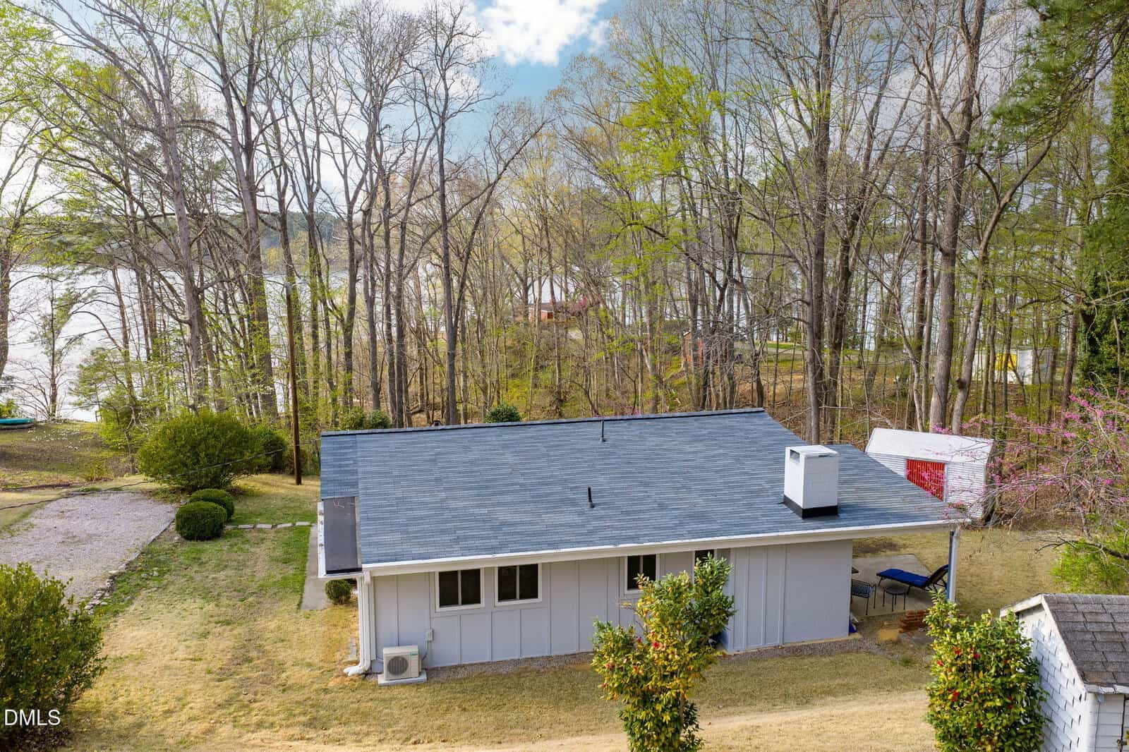 A single-story gray house with a blue roof sits on a grassy lot near a lake, surrounded by tall trees. A small white shed is in the background, and water is visible through the trees.