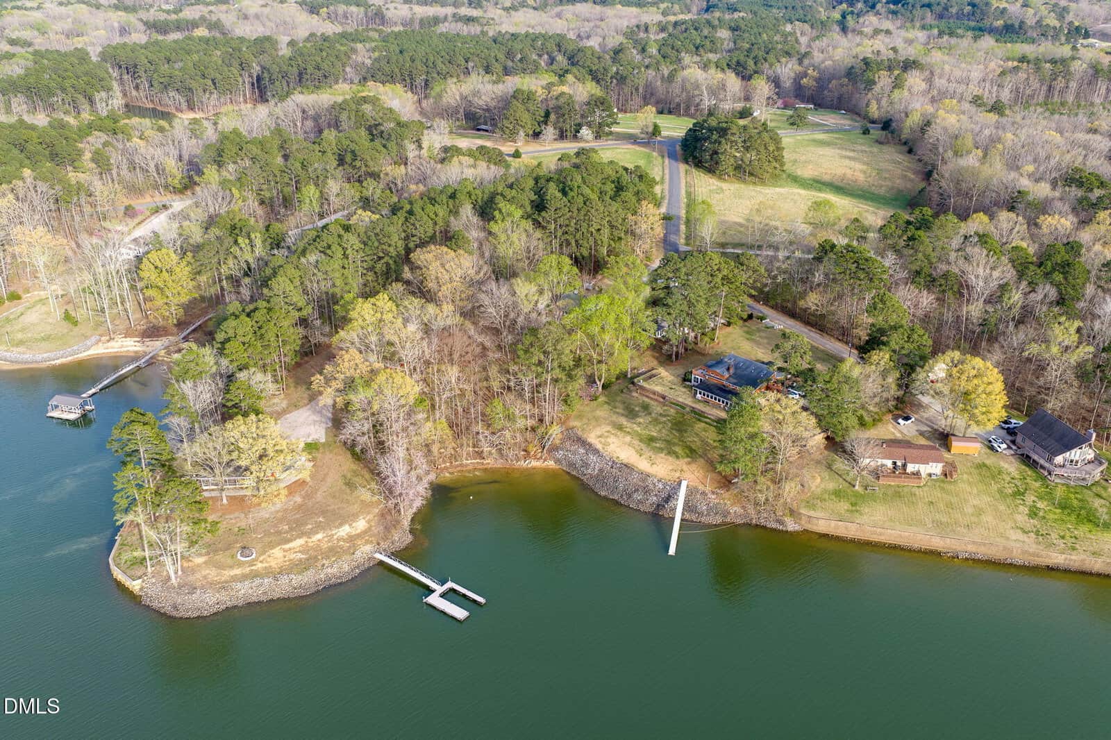 Aerial view of a wooded lakeside area with several houses, docks extending into the water, and grassy clearings. Multiple trees surround the buildings and shoreline, with a road running through the background.