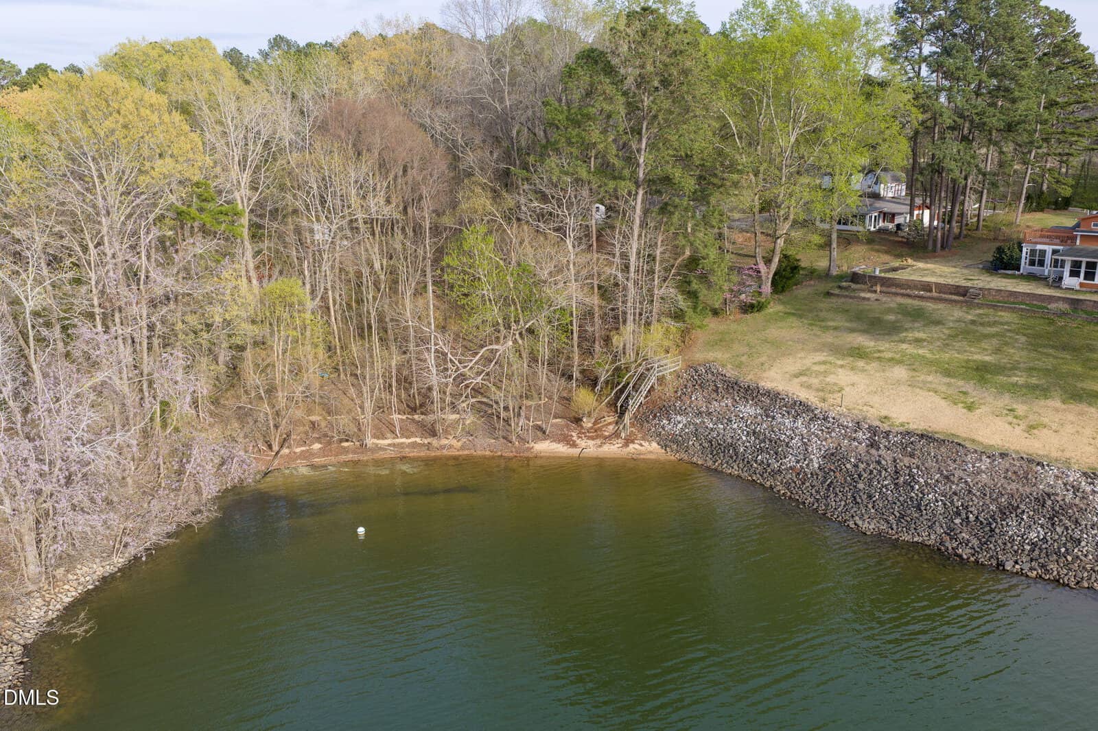 A shoreline with a mix of bare and leafy trees, a rocky embankment, and a small sandy area by the water; houses are visible through the trees on the grassy area above the embankment.