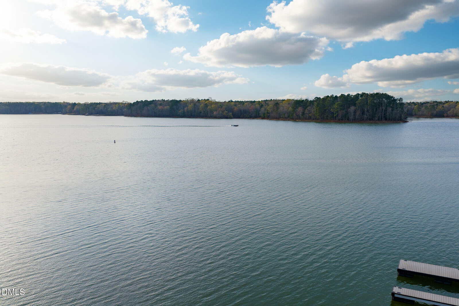 A calm lake with gentle ripples, bordered by a tree-lined shore under a partly cloudy blue sky. A boat is visible in the distance, and parts of docks are seen in the lower right corner.