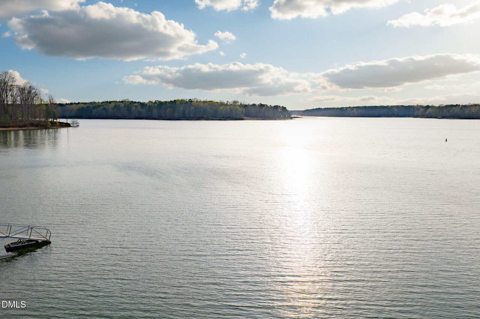 A calm lake under a partly cloudy sky, with sunlight reflecting on the water. Forested shoreline is visible in the distance, and a small dock is at the lower left.