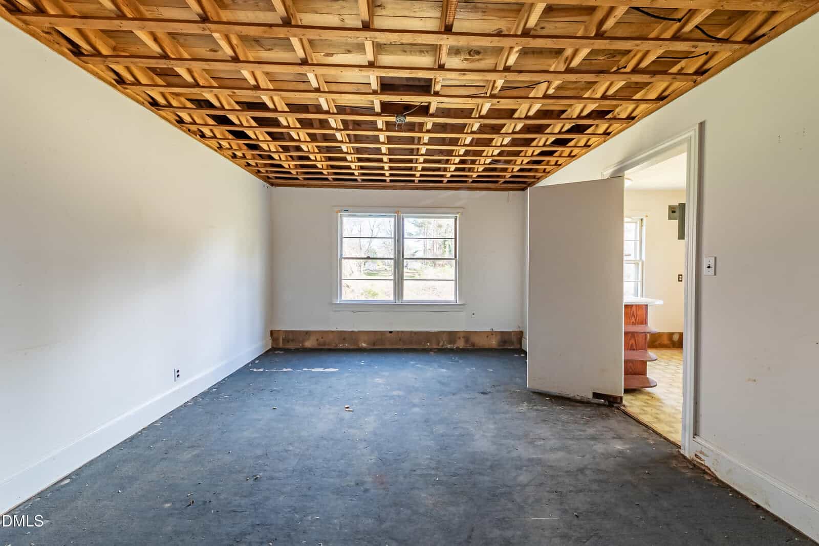 Empty room with white walls, a unique wooden grid ceiling, a blue carpet, baseboard heater, and a large window. An open door leads to another room with visible brown flooring and partial view of kitchen cabinets.