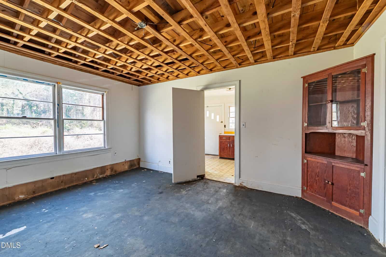 Empty room with exposed wooden ceiling, old dark carpet, and white walls. Large windows on the left, built-in wooden cabinet on the right, and open doorway leading to another room with linoleum flooring.