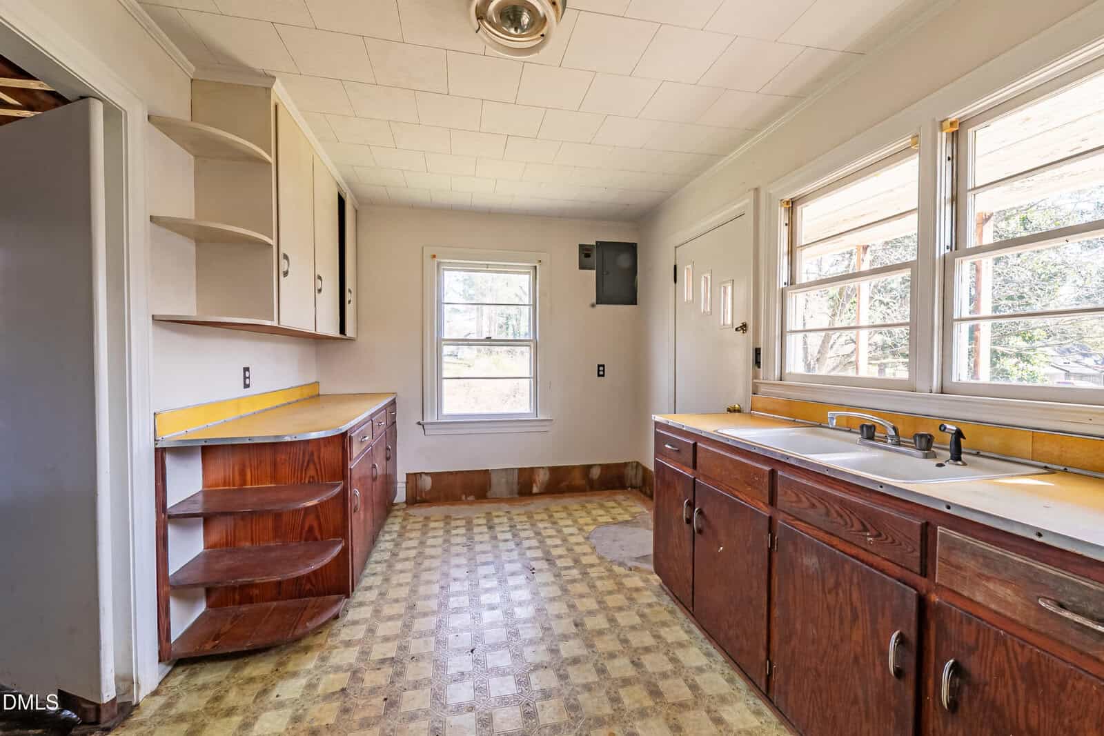 Old kitchen with worn linoleum floor, wooden cabinets, open shelves, double sink under three windows, and a yellowed countertop. Natural light streams in through windows on two walls.