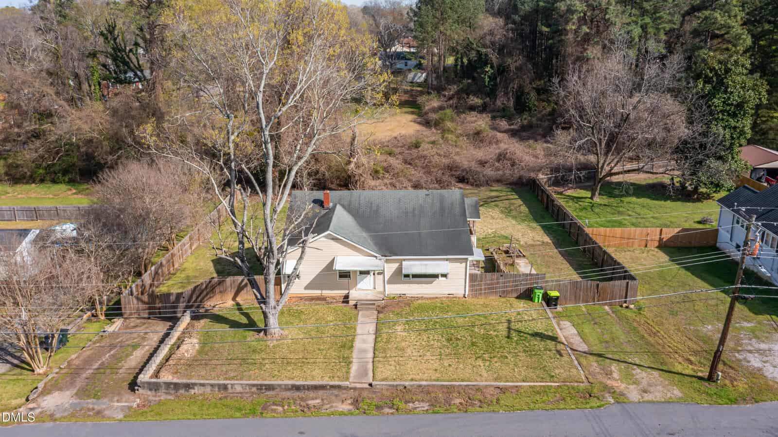 Aerial view of a single-story beige house with a gray roof, large front yard, concrete walkway, and fenced backyard. Leafless trees and neighboring houses are visible, with a wooded area behind the property.