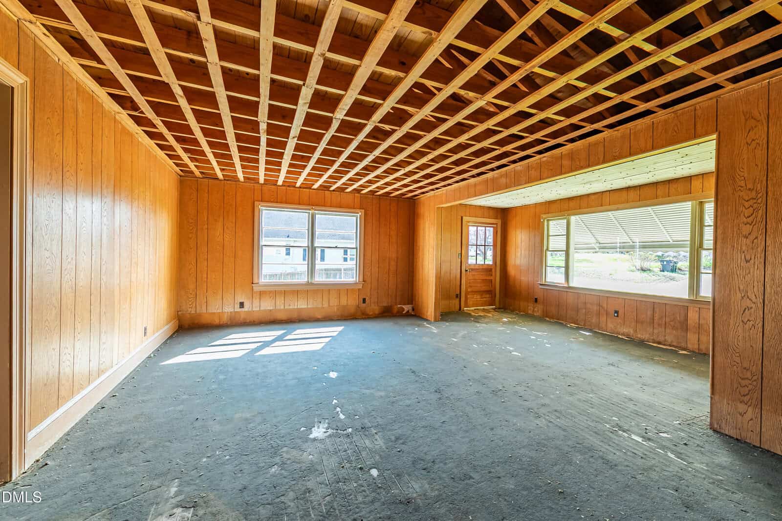 Unfinished room with wood panel walls, exposed grid ceiling, and large windows letting in natural light. A door leads outside, and the floor is bare without any covering.