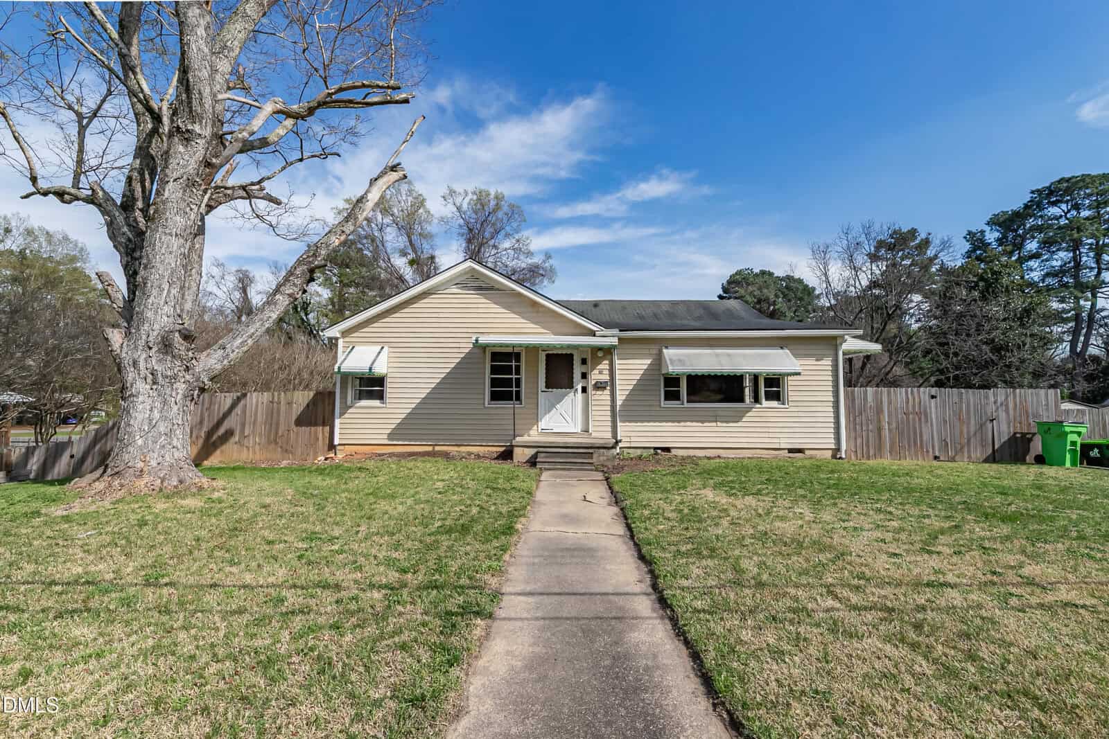 Single-story beige house with a dark roof, white trim, and two awnings over the front windows. A concrete walkway leads to the front door. The yard has grass, a large leafless tree, and is enclosed by a wooden fence.