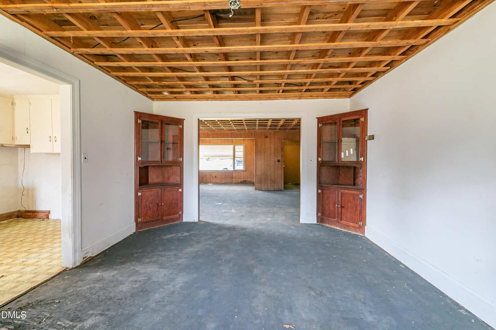 Empty room with dark carpet, white walls, and a wood grid ceiling. Built-in wooden shelves flank double doorways leading to another room. Kitchen with patterned floor is partially visible on the left.