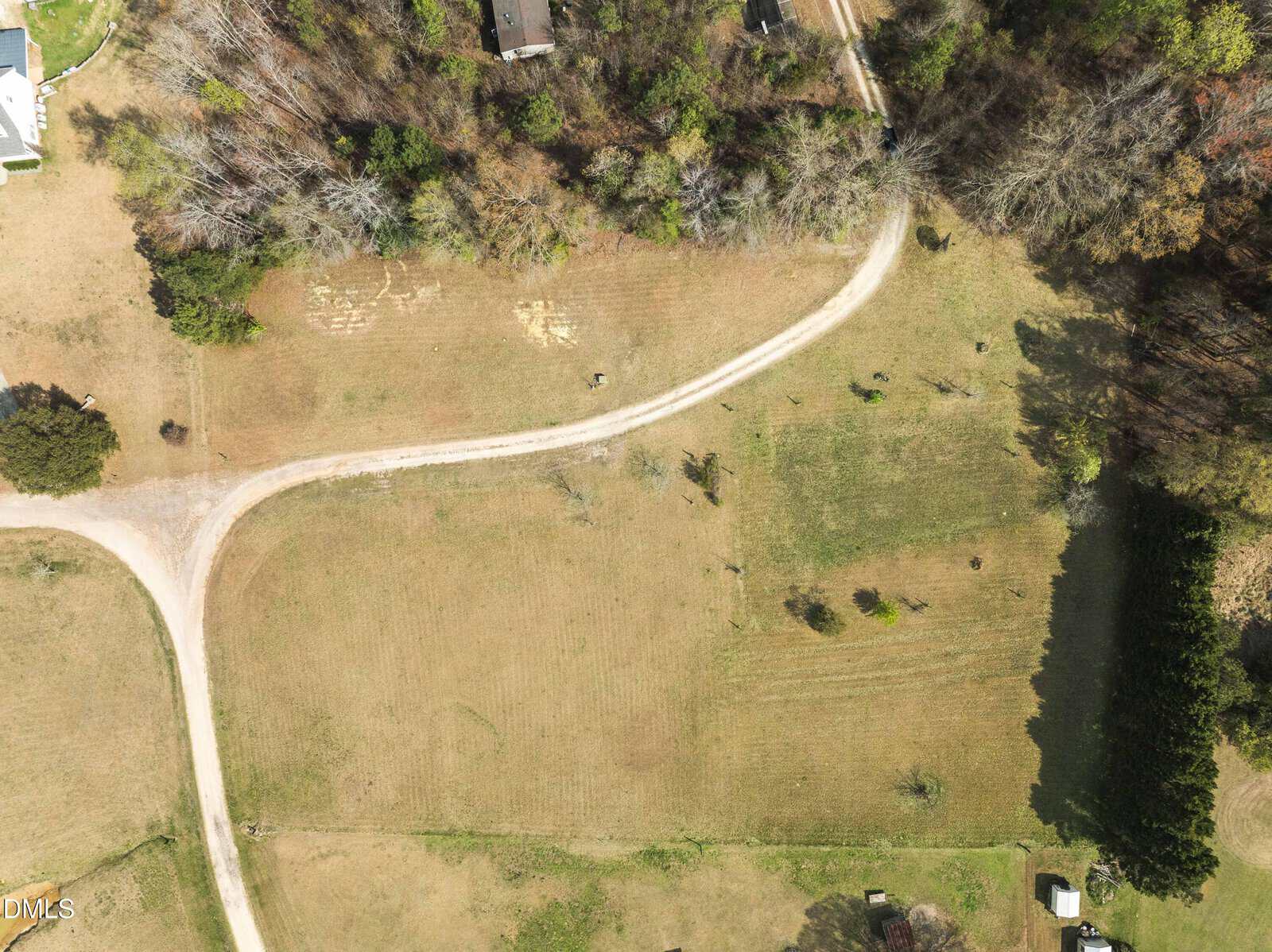 Aerial view of a curved dirt road passing through a grassy area with scattered trees and buildings at the edges. The landscape is mostly open with some patches of trees and shadows.