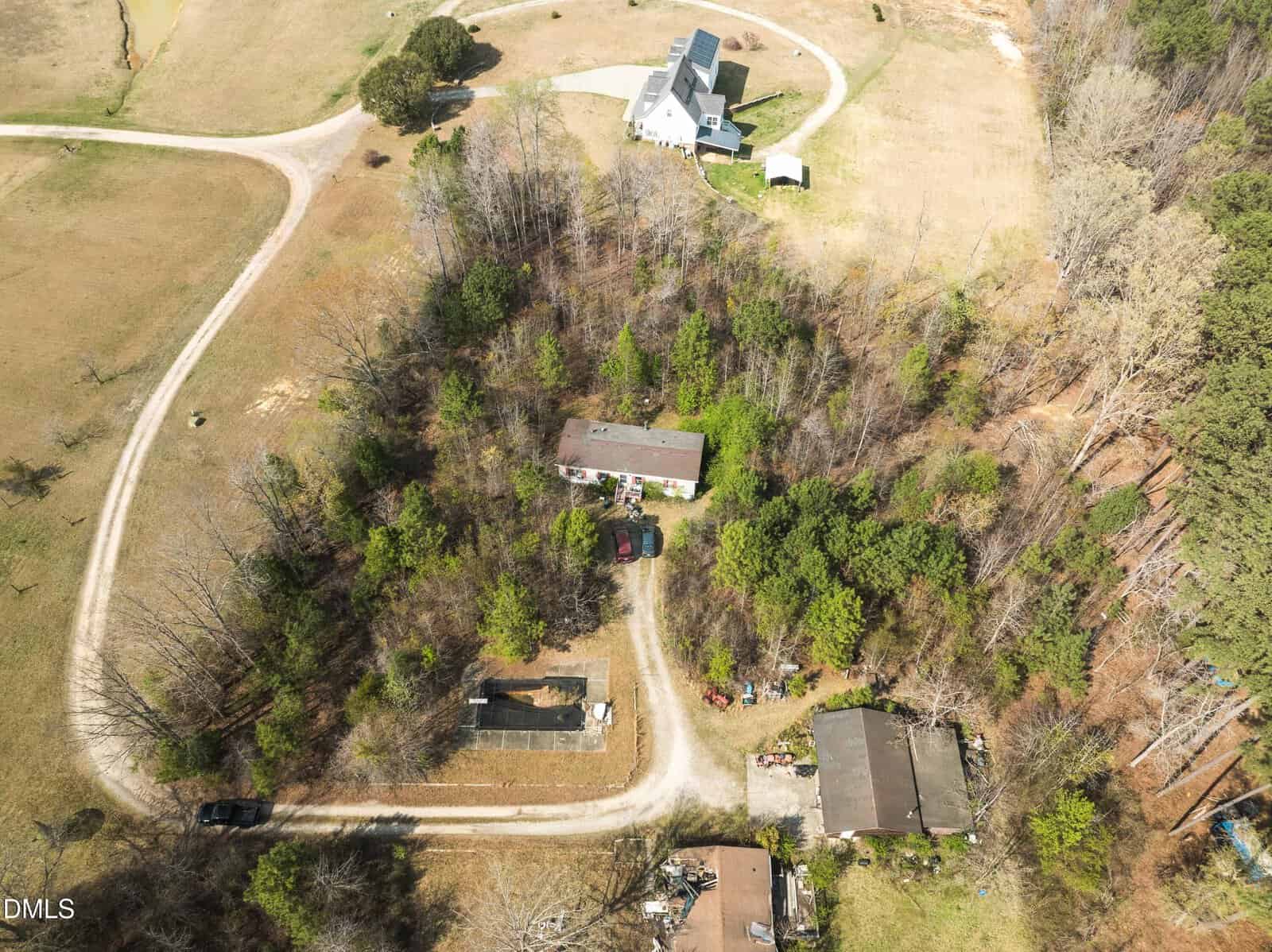 Aerial view of a rural area showing a curved dirt driveway leading to three single-story houses surrounded by trees, with a larger house and open fields in the upper part of the image.